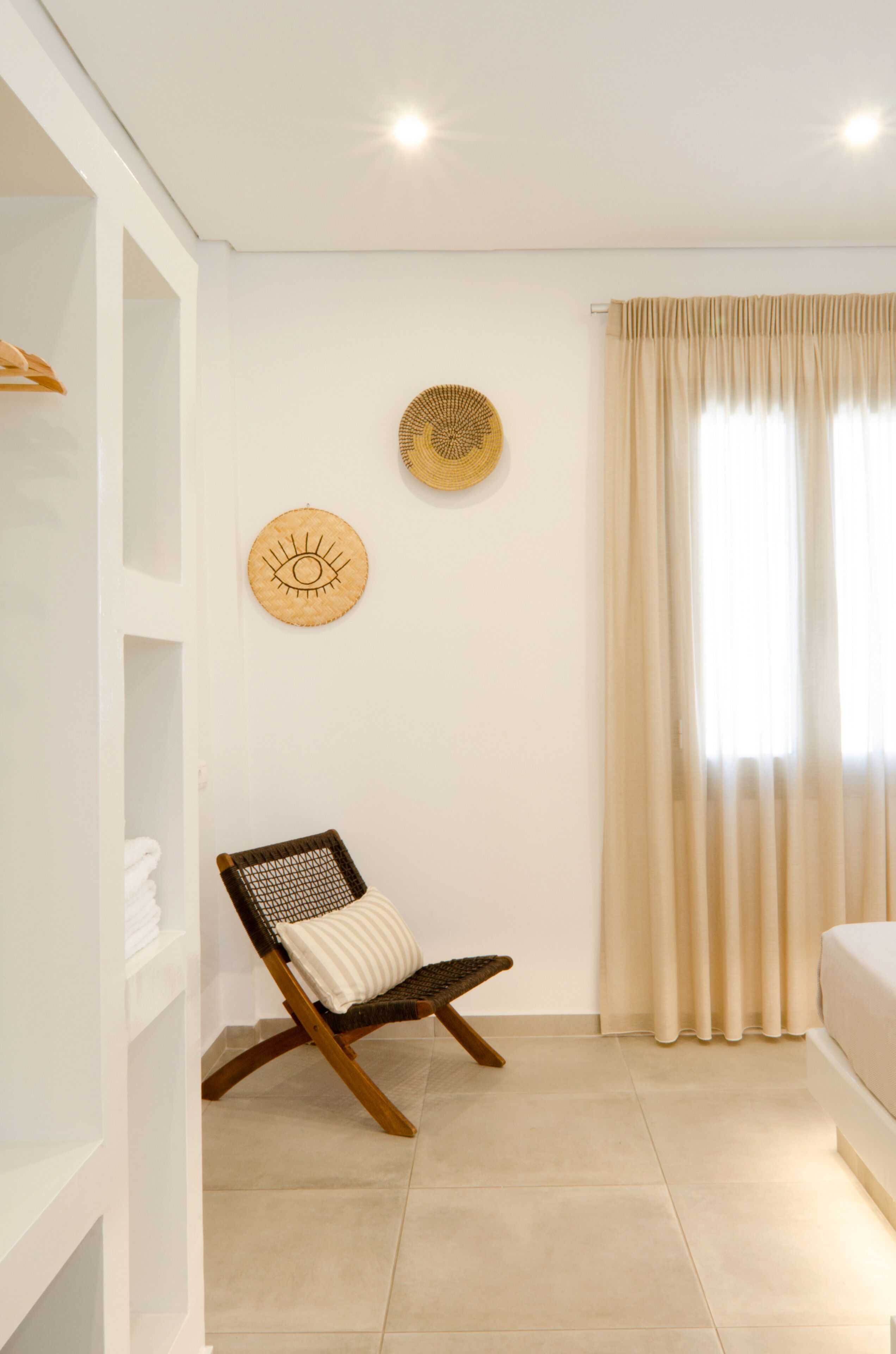Una habitación con pared blanca, una silla de madera con cojín, un estante con toallas dobladas y una ventana con cortinas beige.