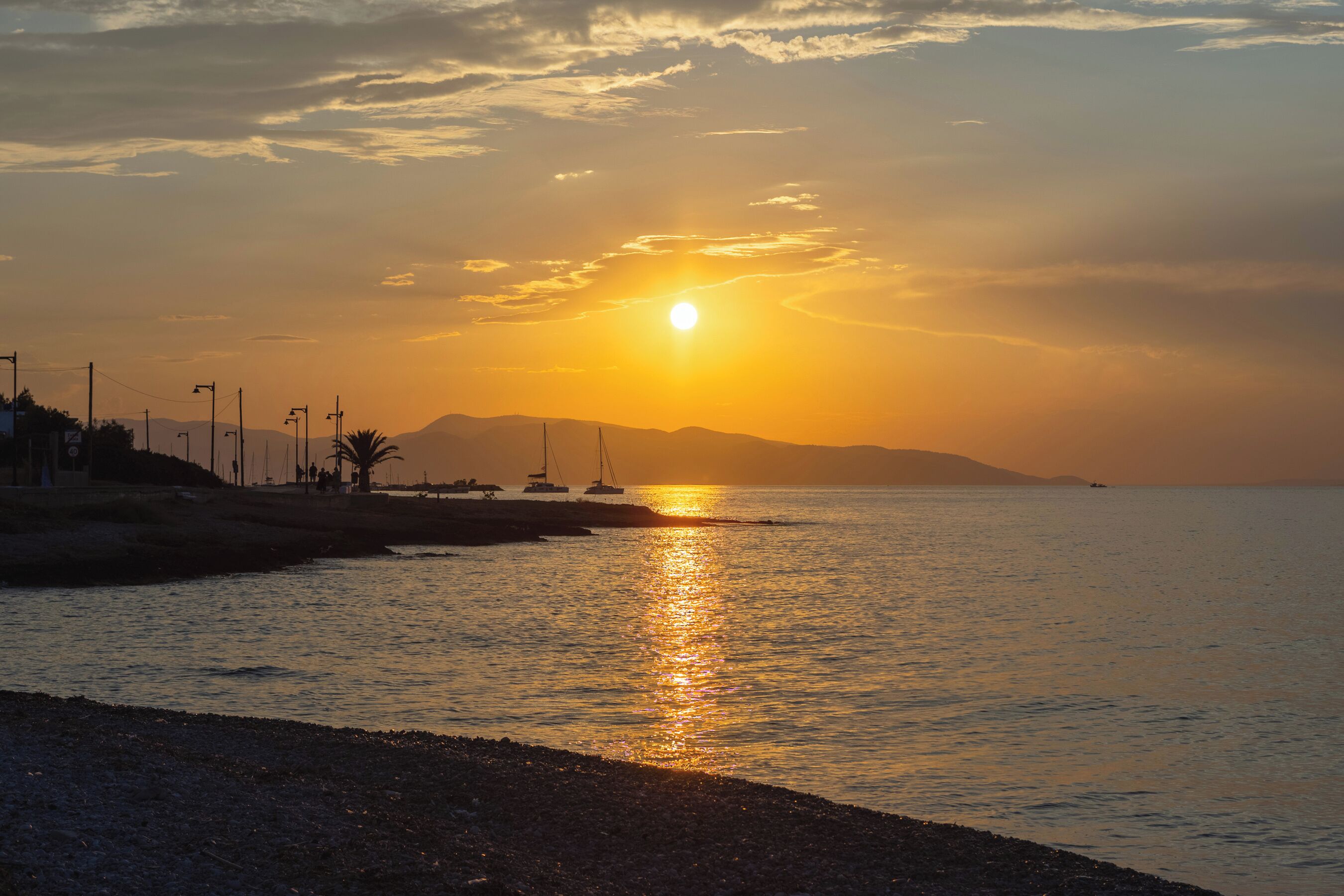 Un coucher de soleil sur une mer calme, avec des bateaux, une plage et des montagnes à l’horizon.
