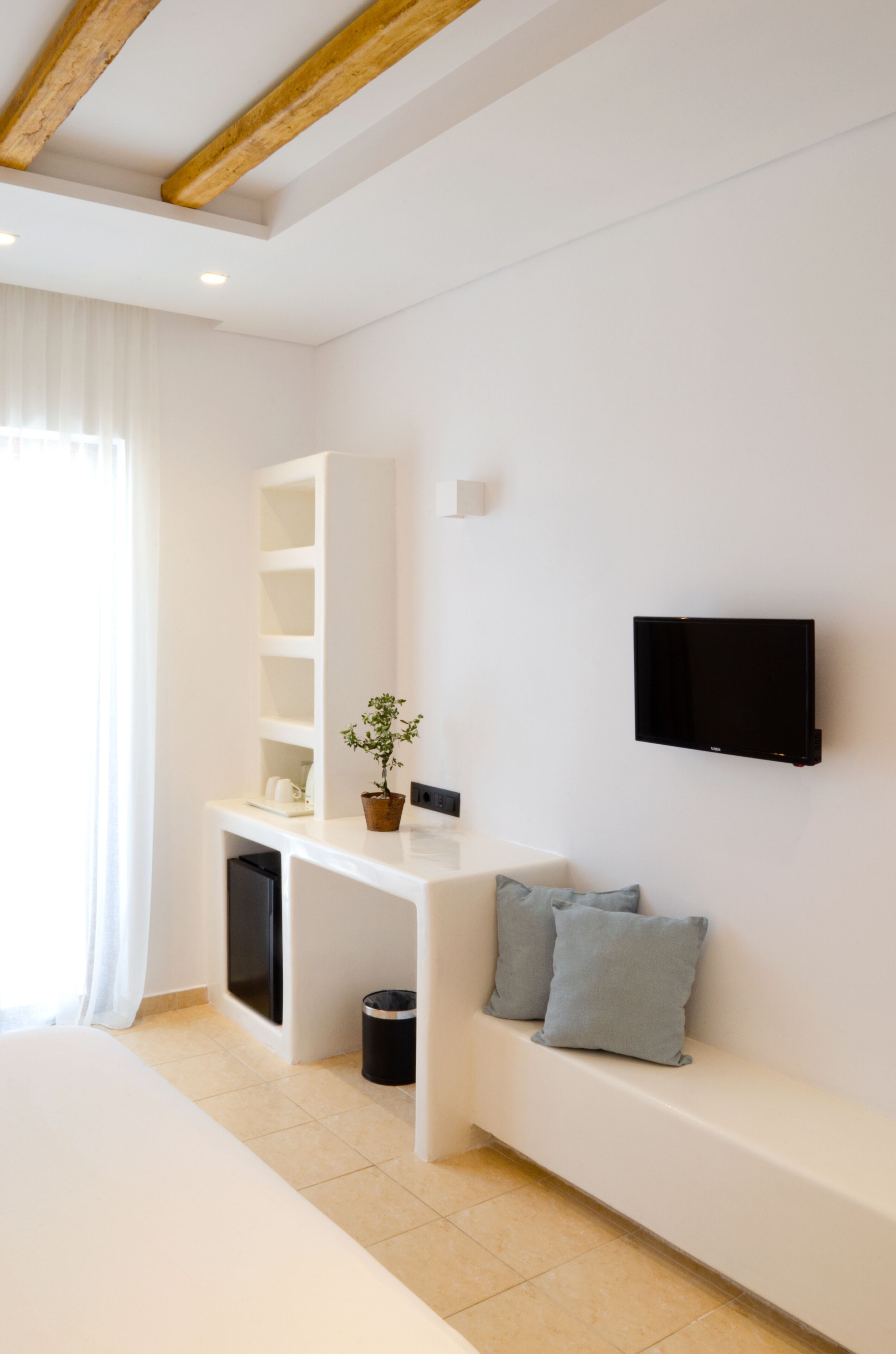 Een moderne hotelkamer met een wit bed, een ingebouwde bank met kussens, een klein tafeltje met een bloempot, een wandgemonteerde televisie en een groot raam met dunne gordijnen.