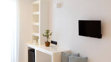 Een moderne hotelkamer met een wit bed, een ingebouwde bank met kussens, een klein tafeltje met een bloempot, een wandgemonteerde televisie en een groot raam met dunne gordijnen.