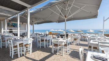 An outdoor dining area with white tables and chairs, umbrellas, and a view of the sea.