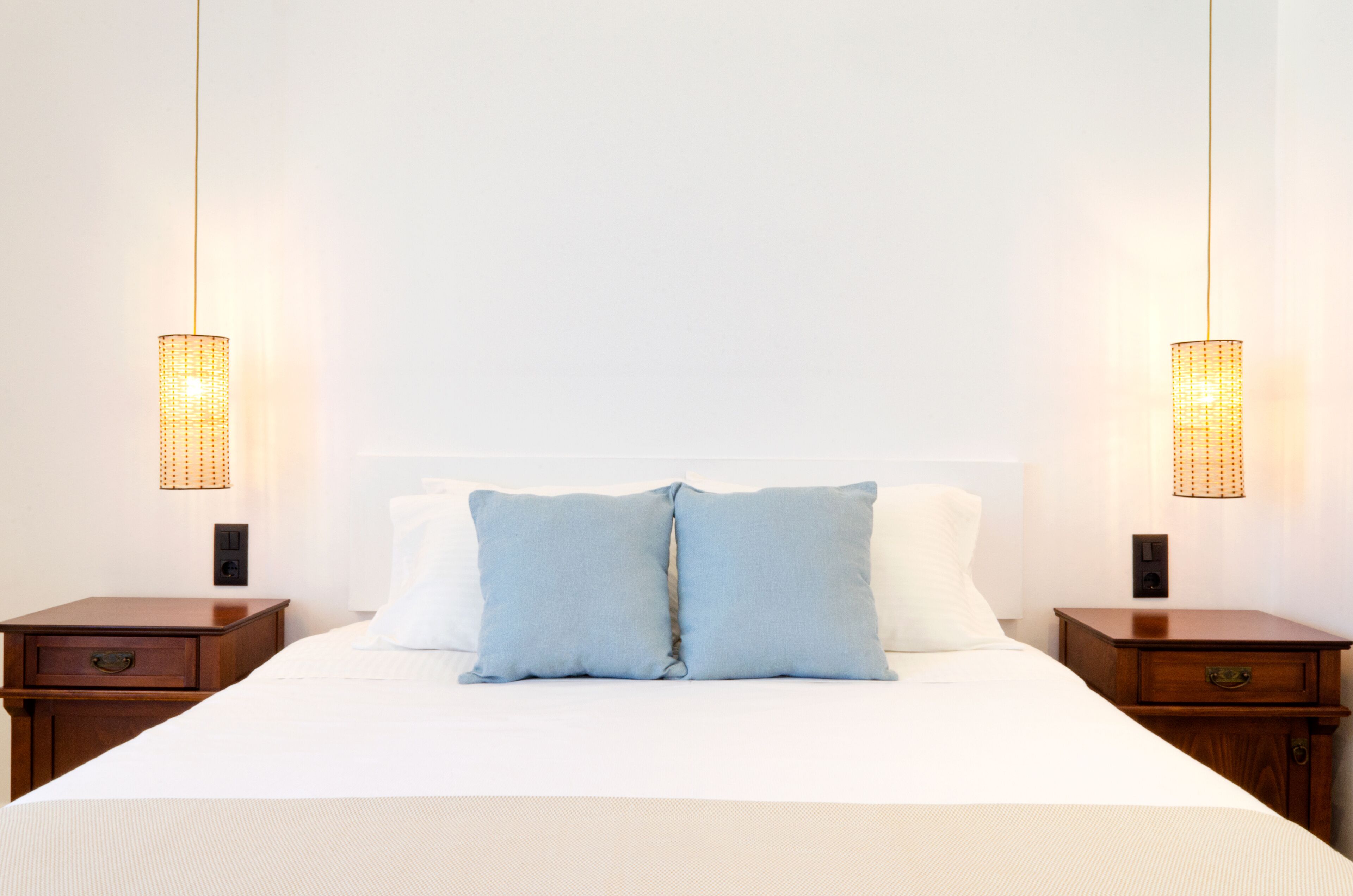 Una cama cuidadosamente tendida con dos almohadas, una mesita de noche de madera con cajón y un aplique de pared a cada lado.