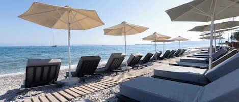A beachside lounge area with sun loungers and umbrellas, a pebble beach, and the sea in the background.