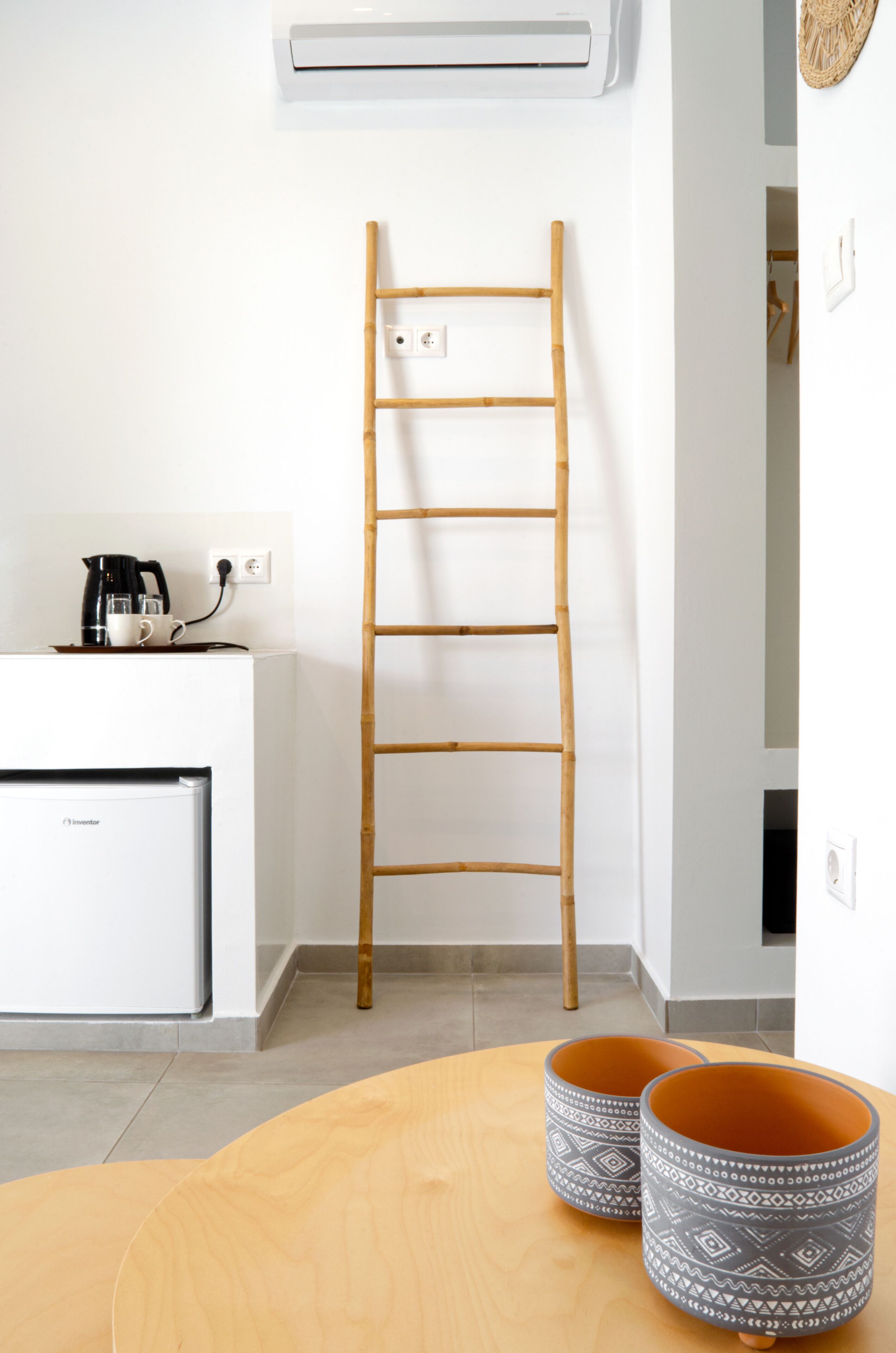 A room with a white wall, a wooden ladder, a small white refrigerator, a coffee maker, and two patterned cushions on a wooden table.