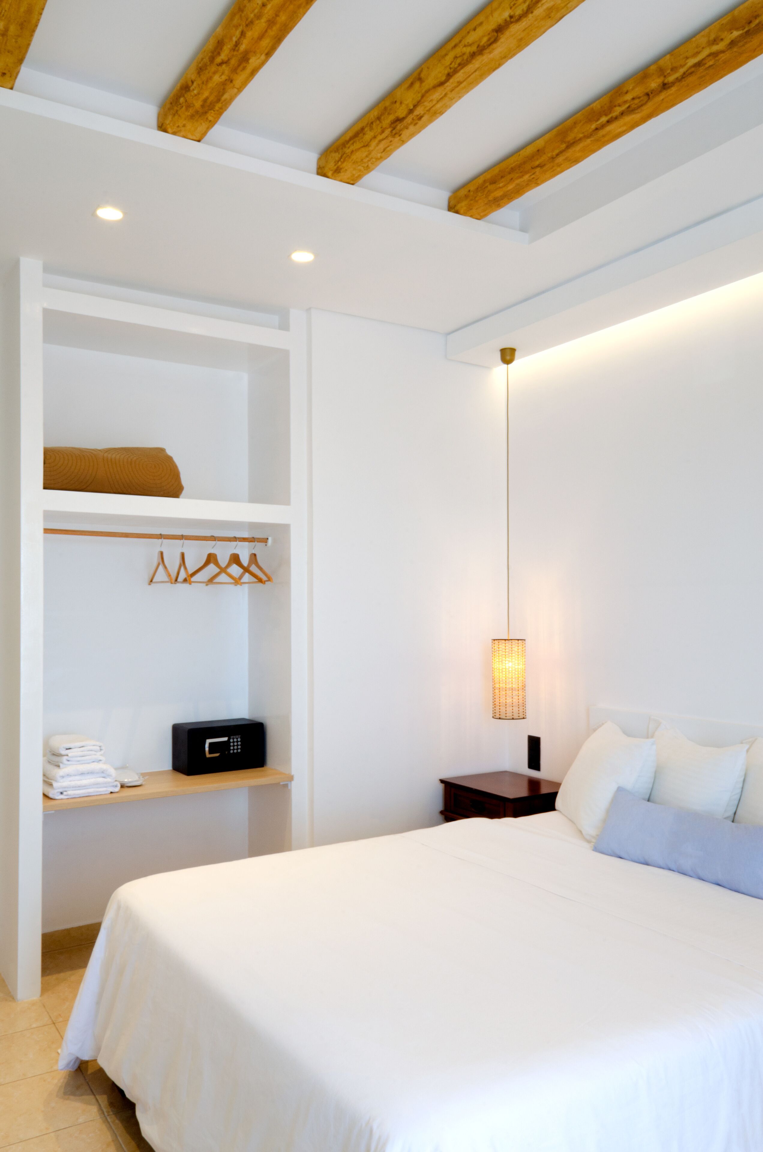 A bedroom with a large bed, wooden beams on the ceiling, a bedside table, a hanging light fixture, and a built-in wardrobe with a hanging rack.