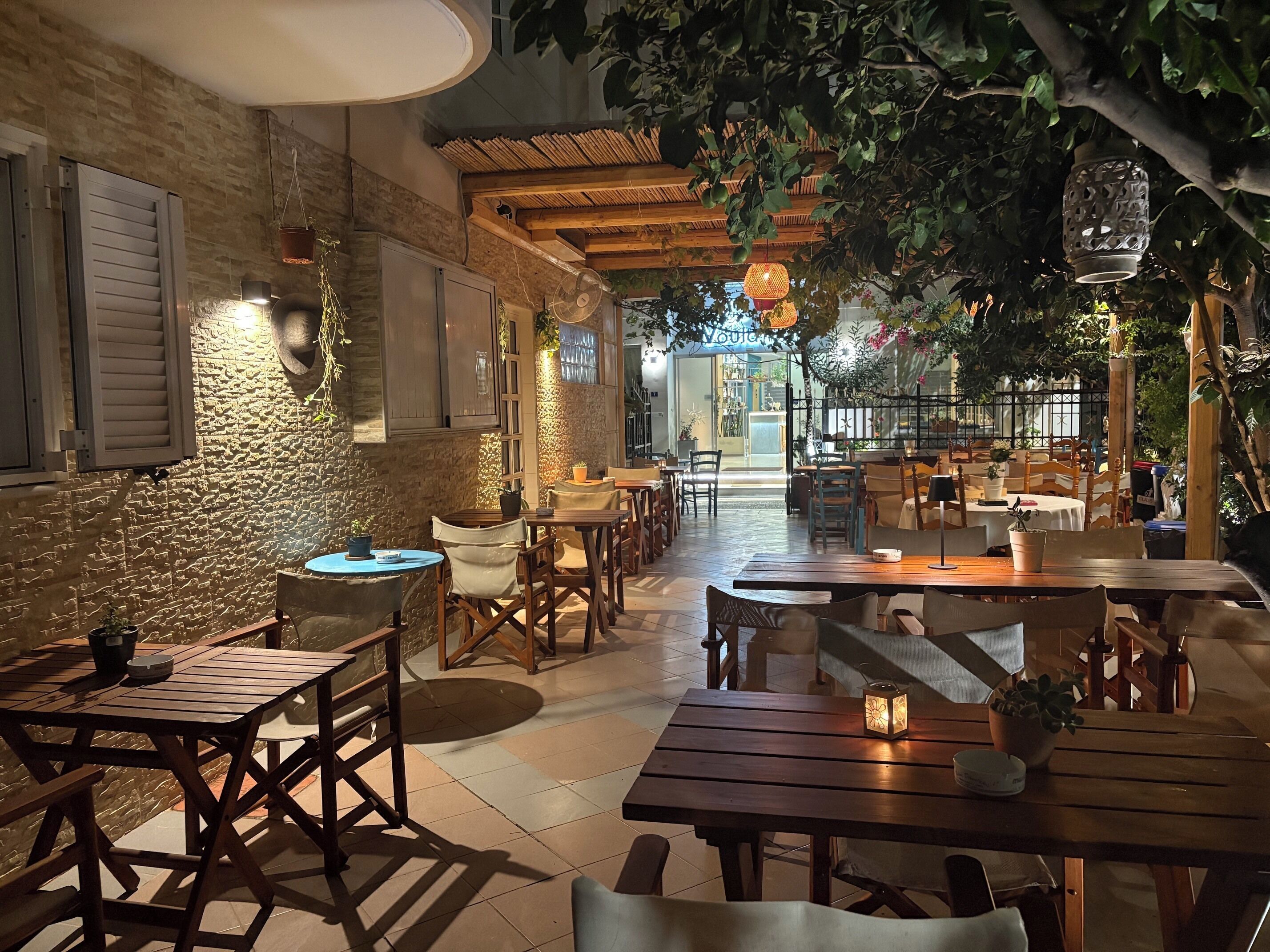 An outdoor dining area with wooden tables and chairs, stone walls, and hanging plants.