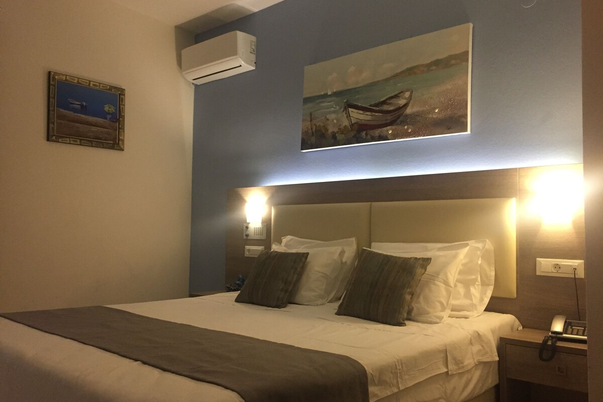 Habitació d'hotel amb llit, làmpades de nit, un quadre d'una embarcació a la paret i un aparell d'aire condicionat.