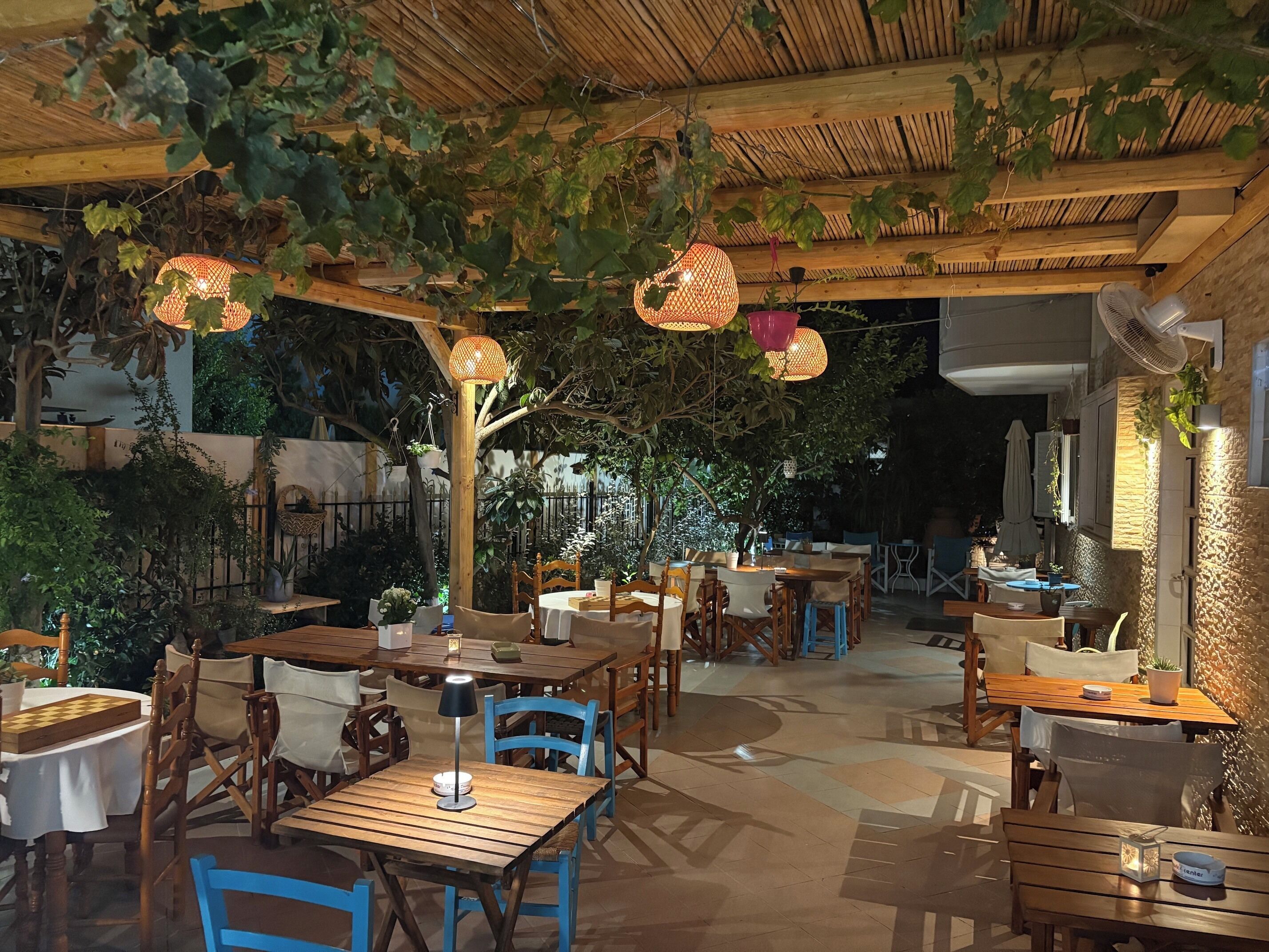 Un área de comedor al aire libre con mesas y sillas de madera, lámparas colgantes y plantas en macetas.