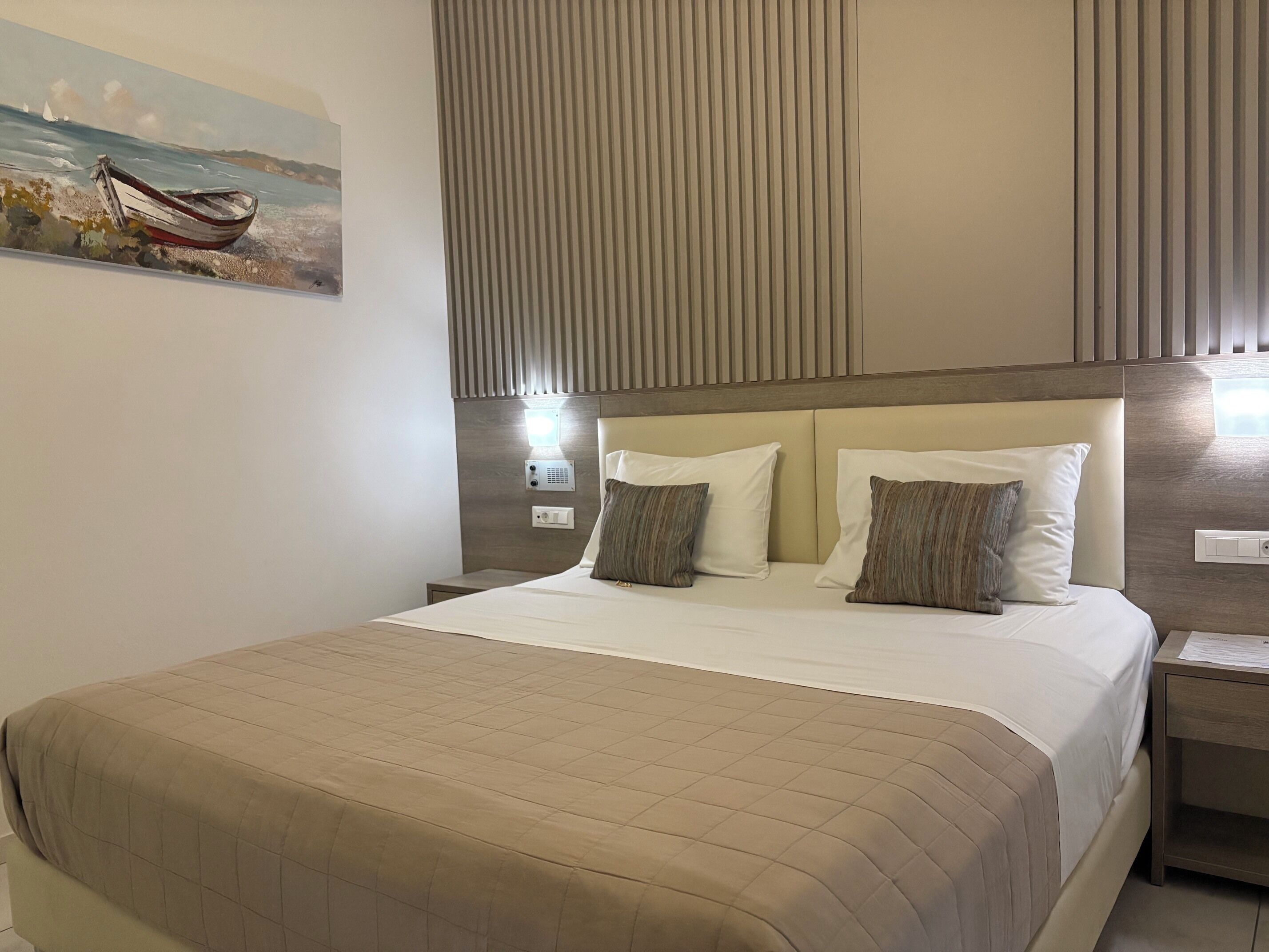 A hotel room with a bed, a nightstand, a wall-mounted light, and a painting of a boat on the wall.