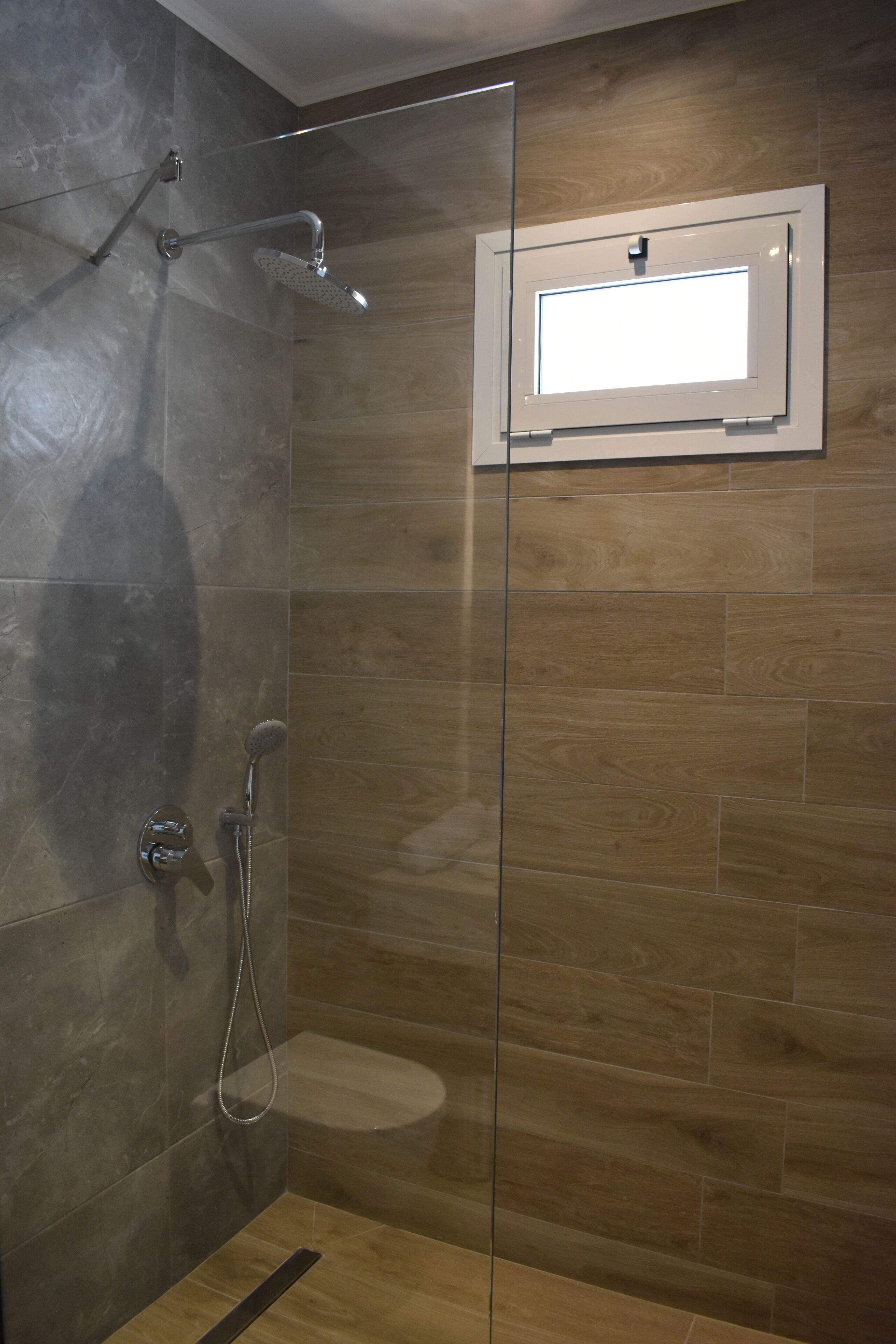 A glass-enclosed shower with a wooden floor and a window above the toilet.