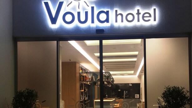 The entrance of Voula Hotel with a modern design, featuring a glass facade, a reception desk, and two potted plants. - Voula Hotel (Hersonissos)