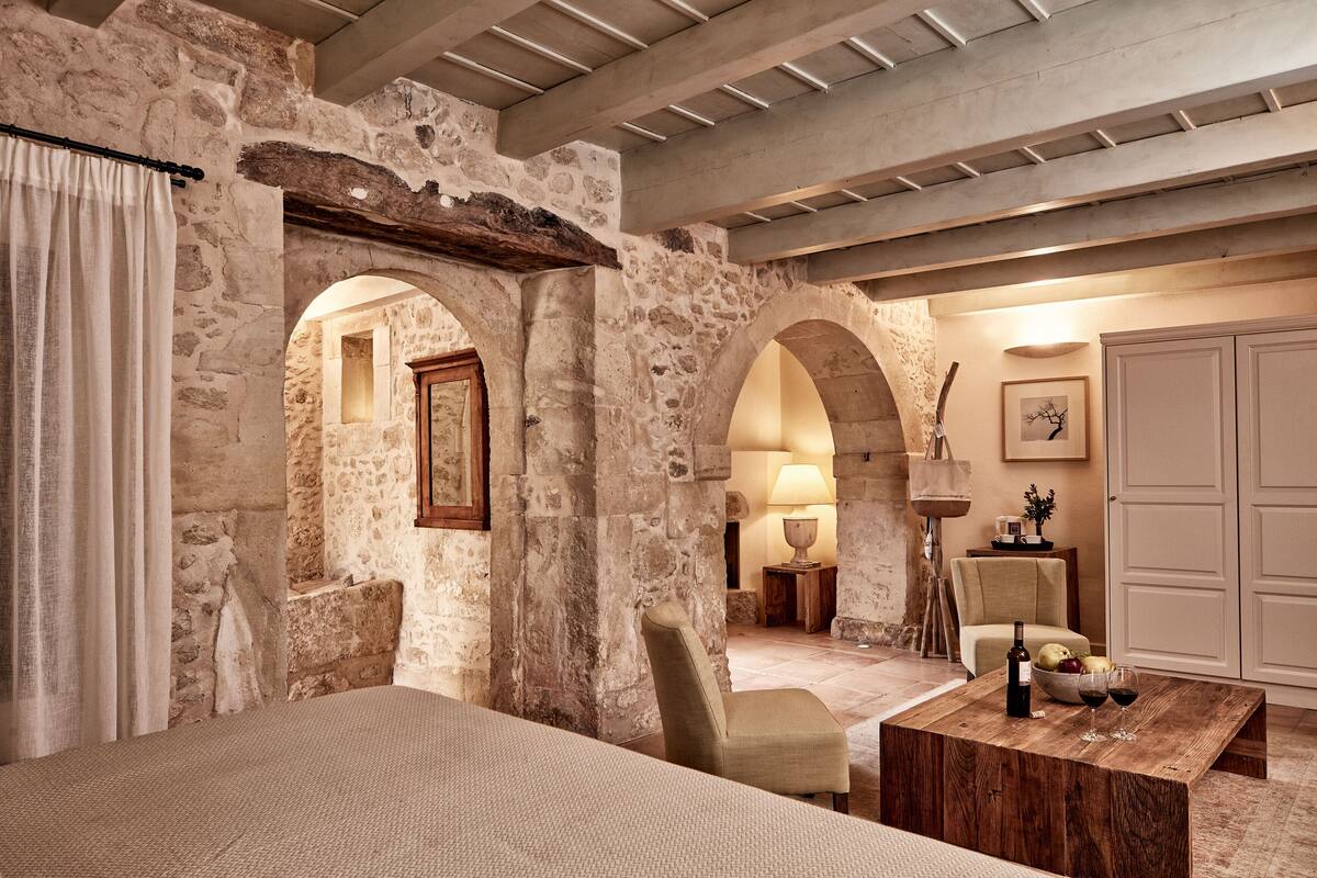 A room with stone walls, a wooden bed, a chair, a table with a bottle and glasses, and a lamp.