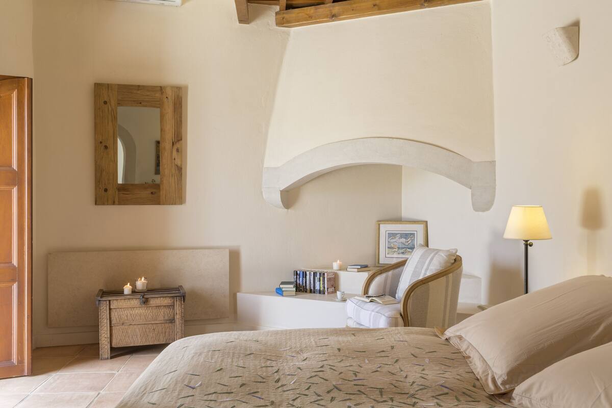 A bedroom with a bed, bedside table, lamp, chair, and a mirror on the wall.