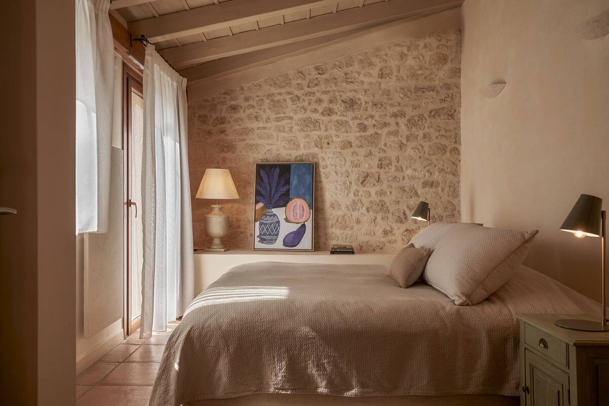 A bedroom with a stone wall, a bed with a beige bedspread, two bedside lamps, and a framed artwork on the wall.