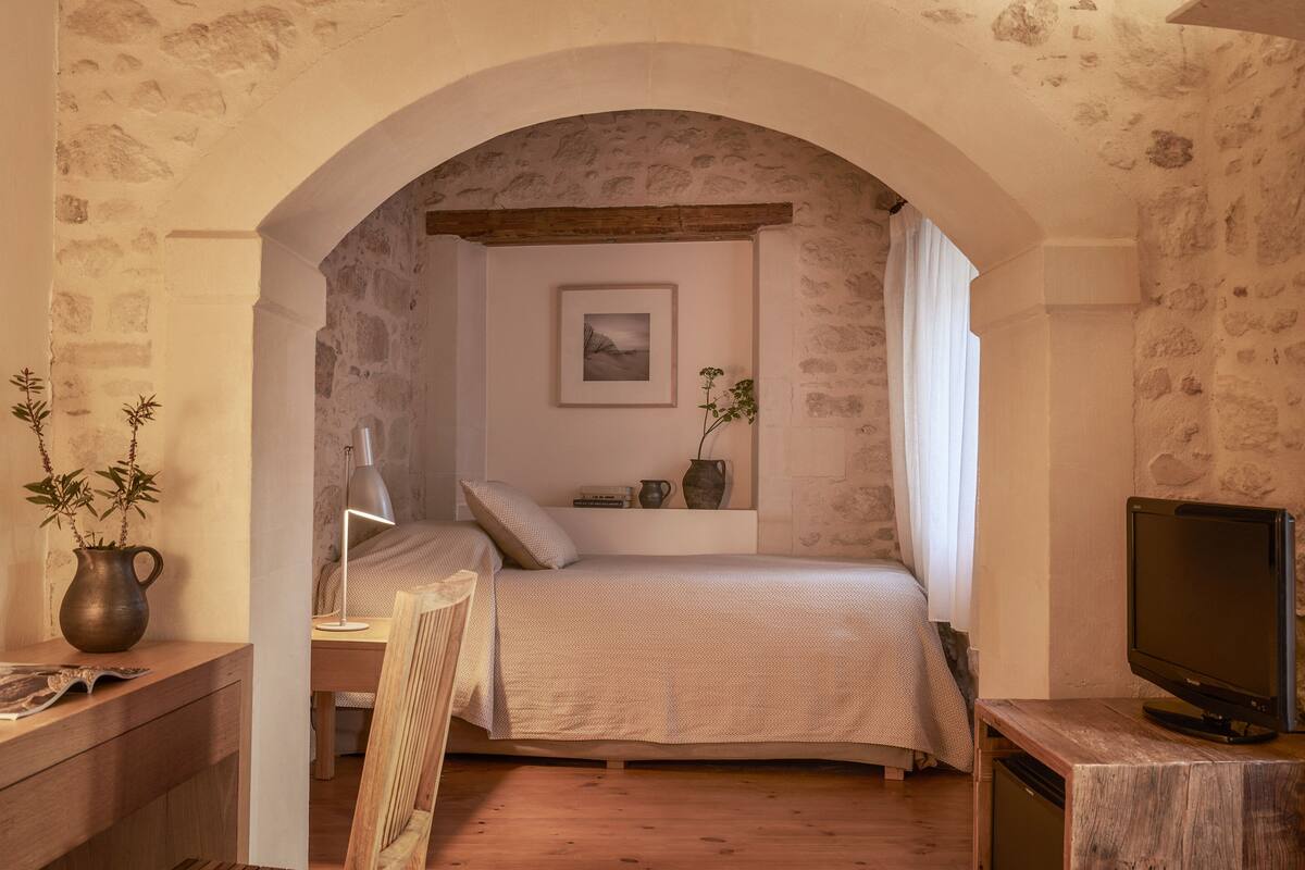 A cozy bedroom with a stone wall, a bed, a desk, a chair, a TV, and a window with curtains.