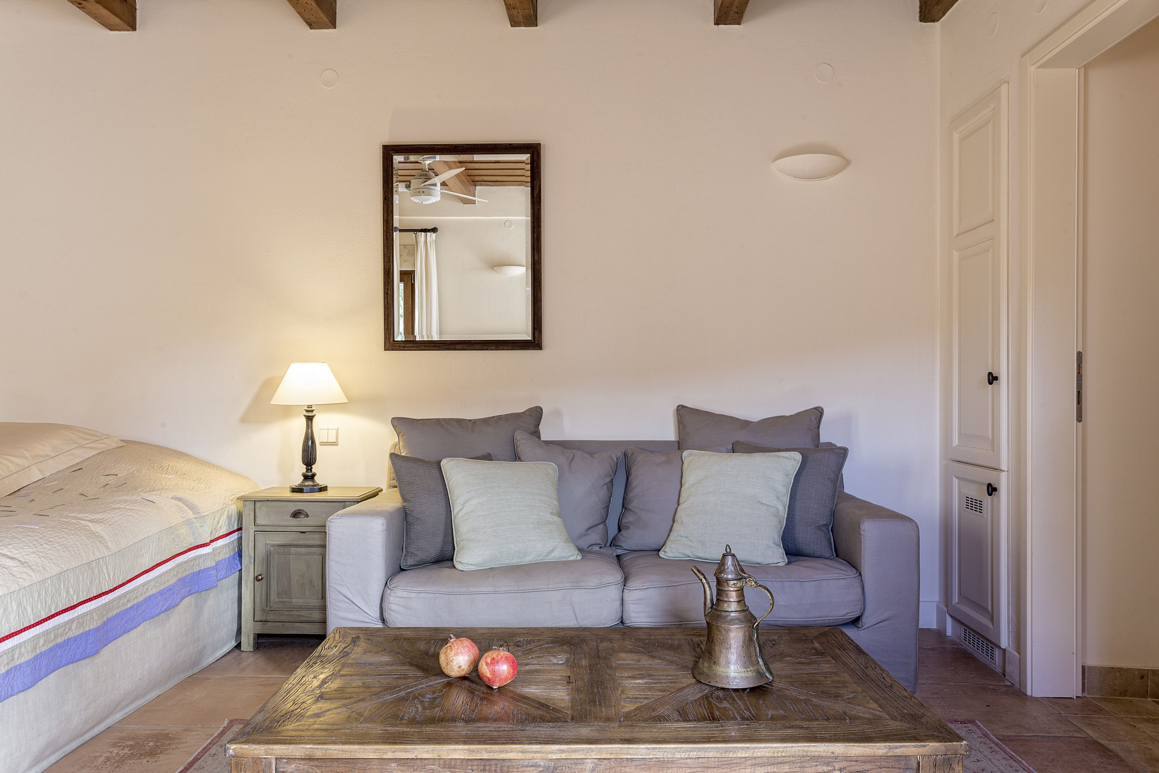 Un dormitorio con una cama, una mesita de noche con lámpara, un sofá con cojines, una mesa de centro con manzanas y una jarra de metal, y una puerta.