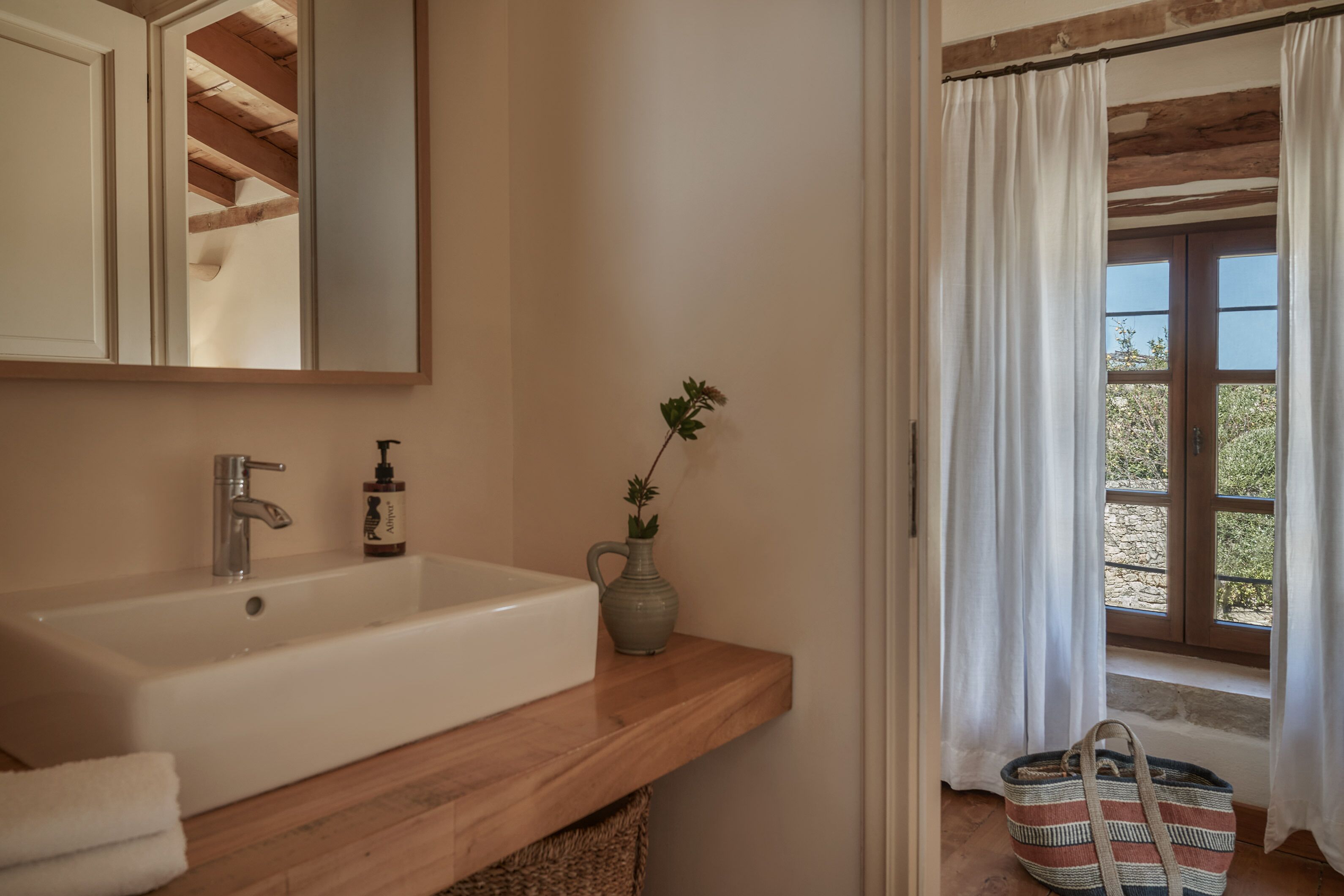 Une salle de bain avec un meuble-lavabo en bois, un lavabo blanc, un miroir, une petite plante en pot et une fenêtre avec des rideaux.