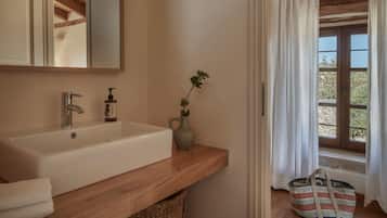 Une salle de bain avec un meuble-lavabo en bois, un lavabo blanc, un miroir, une petite plante en pot et une fenêtre avec des rideaux.