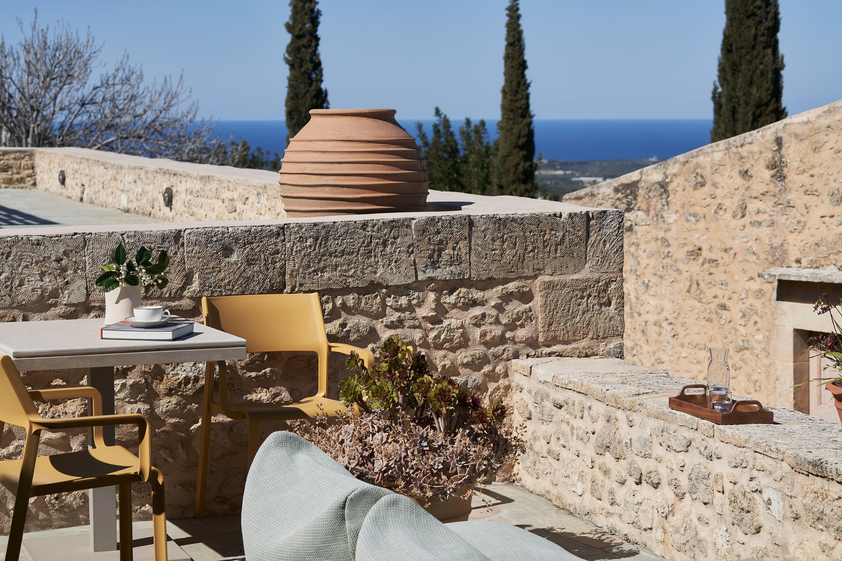A stone wall with a large terracotta pot, a table with a book and a cup, and a chair.
