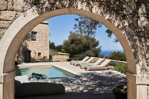 An outdoor swimming pool with lounge chairs and a stone building in the background. - Kapsaliana Village Hotel (Rethymno)