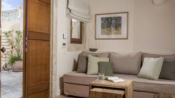 Ein Wohnzimmer mit einem beigefarbenen Sofa, einem Holztisch, einem Fenster mit weißem Rollo, einem gerahmten Bild an der Wand und einem Blumentopf.