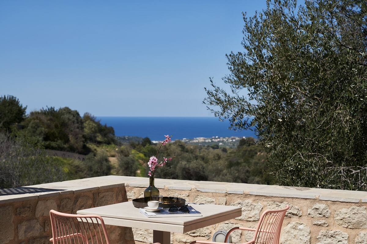 A table with a vase of flowers, a bowl, and a pair of sunglasses on a terrace overlooking a coastal landscape.