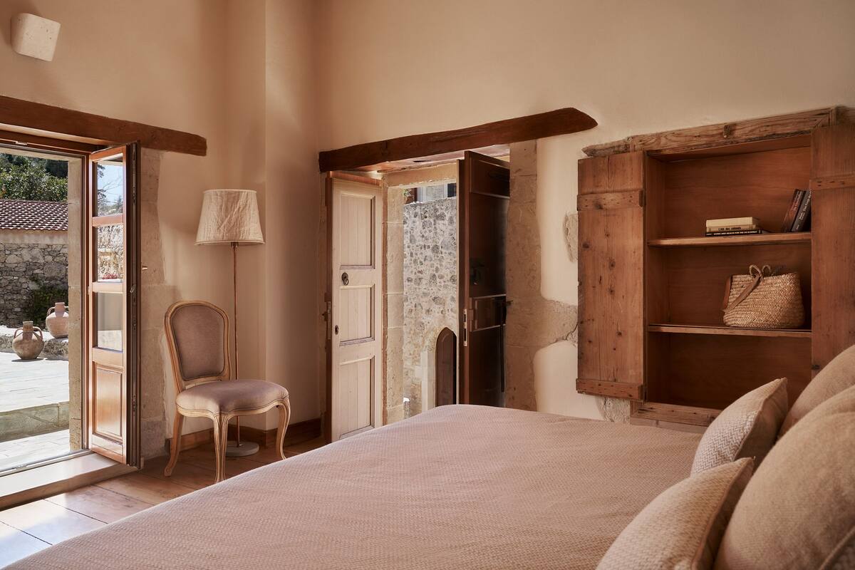 A bedroom with a bed, a chair, a wooden shelf, and a door leading to an outdoor area.