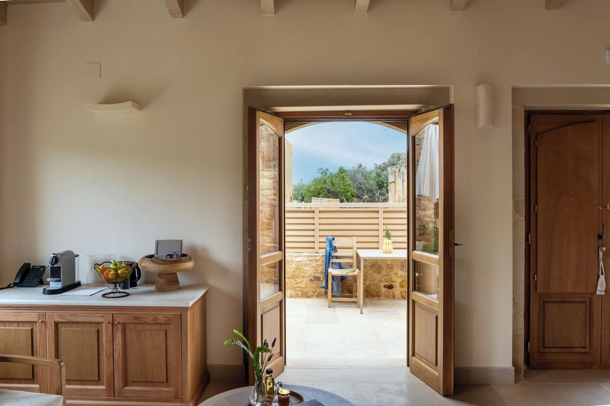 A room with a wooden desk, a telephone, a fruit bowl, and a view of an outdoor patio with a stone wall and a wooden fence.