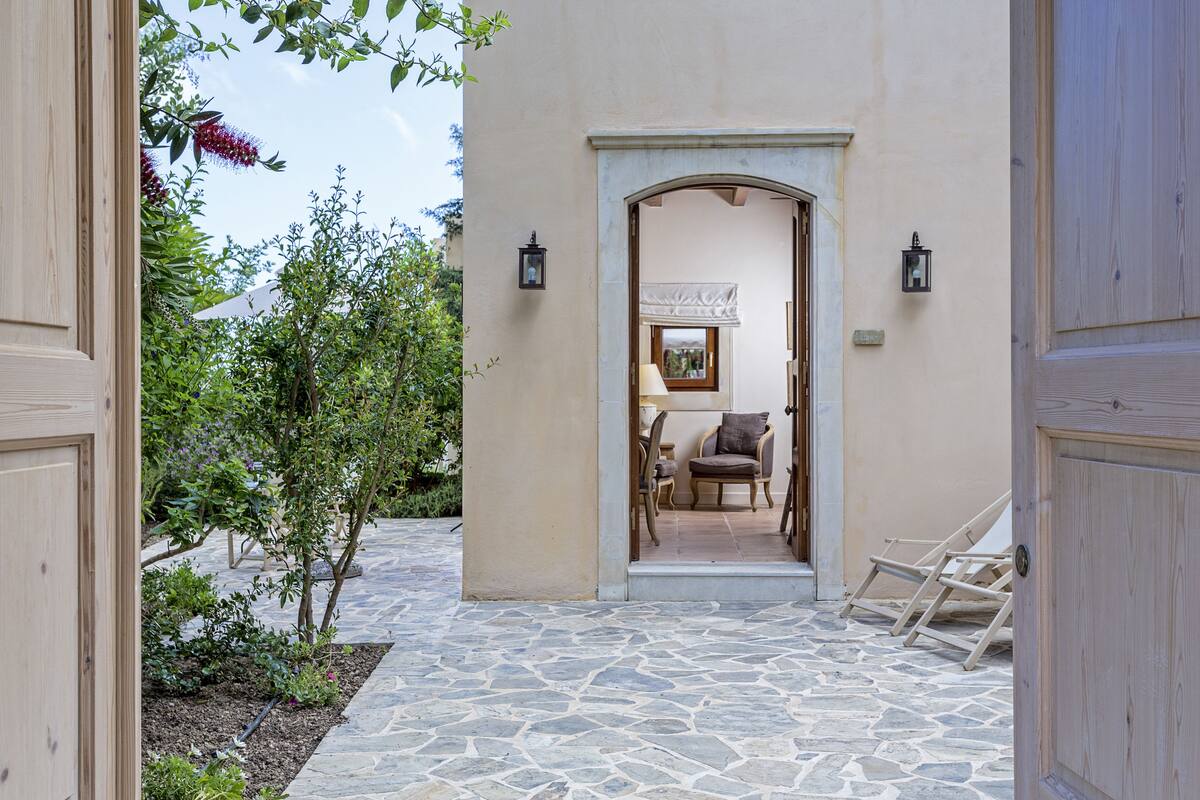 A stone-paved entrance to a building with a wooden door and a small seating area with a chair and a table.