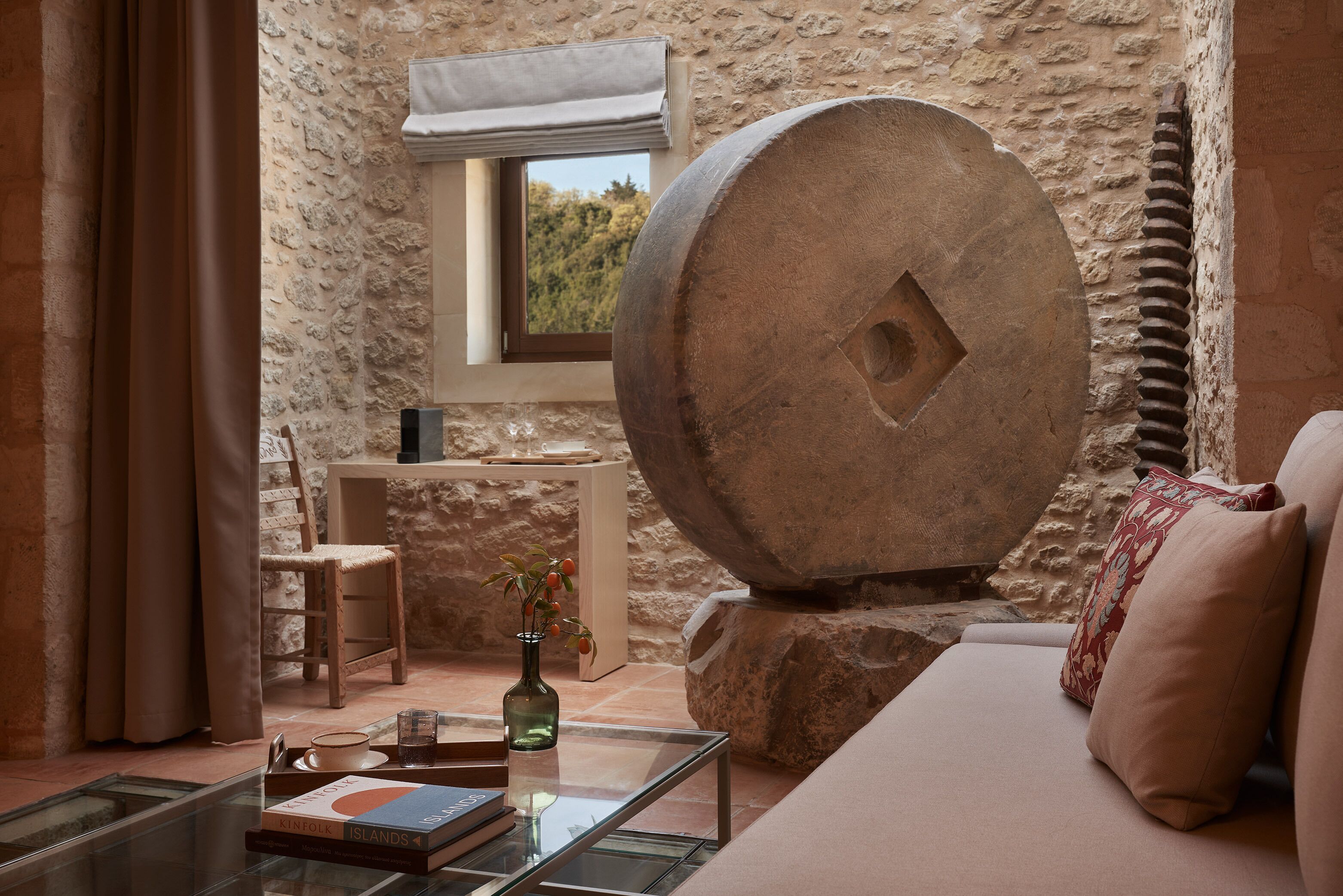 A stone-walled room with a large stone sculpture, a window with a view, a sofa, and a small table with books and a vase.
