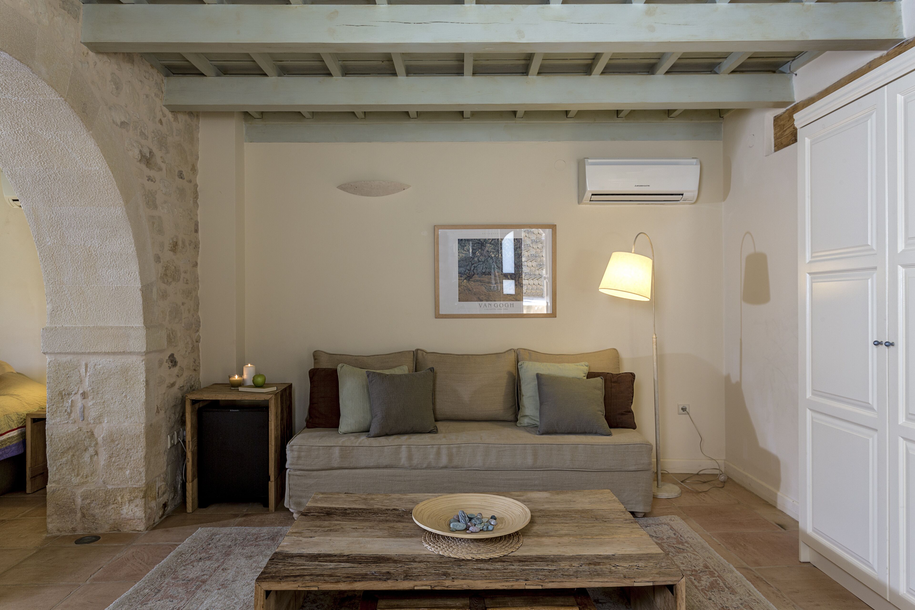 Una sala de estar acogedora con un pilar de piedra, una mesa de centro de madera con un cuenco, un sofá con cojines, una lámpara de pie y un armario blanco.