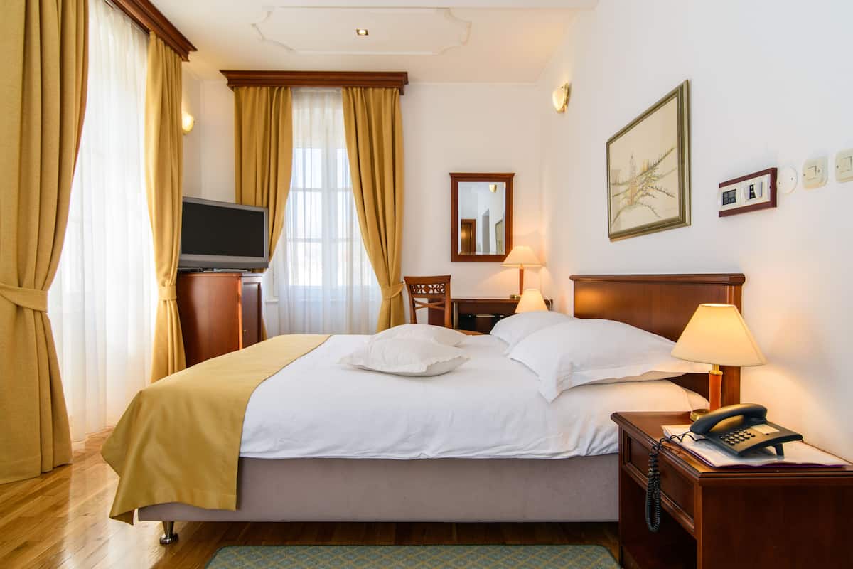 A hotel room with a bed, a TV, a desk with a lamp, a telephone, and a painting on the wall.