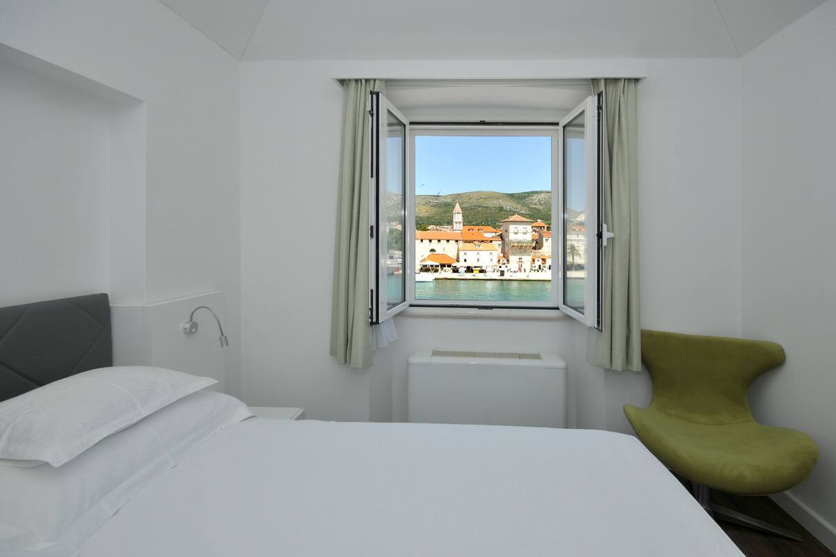 A hotel room with a bed, a green chair, a window with a view of buildings and hills, and a radiator.