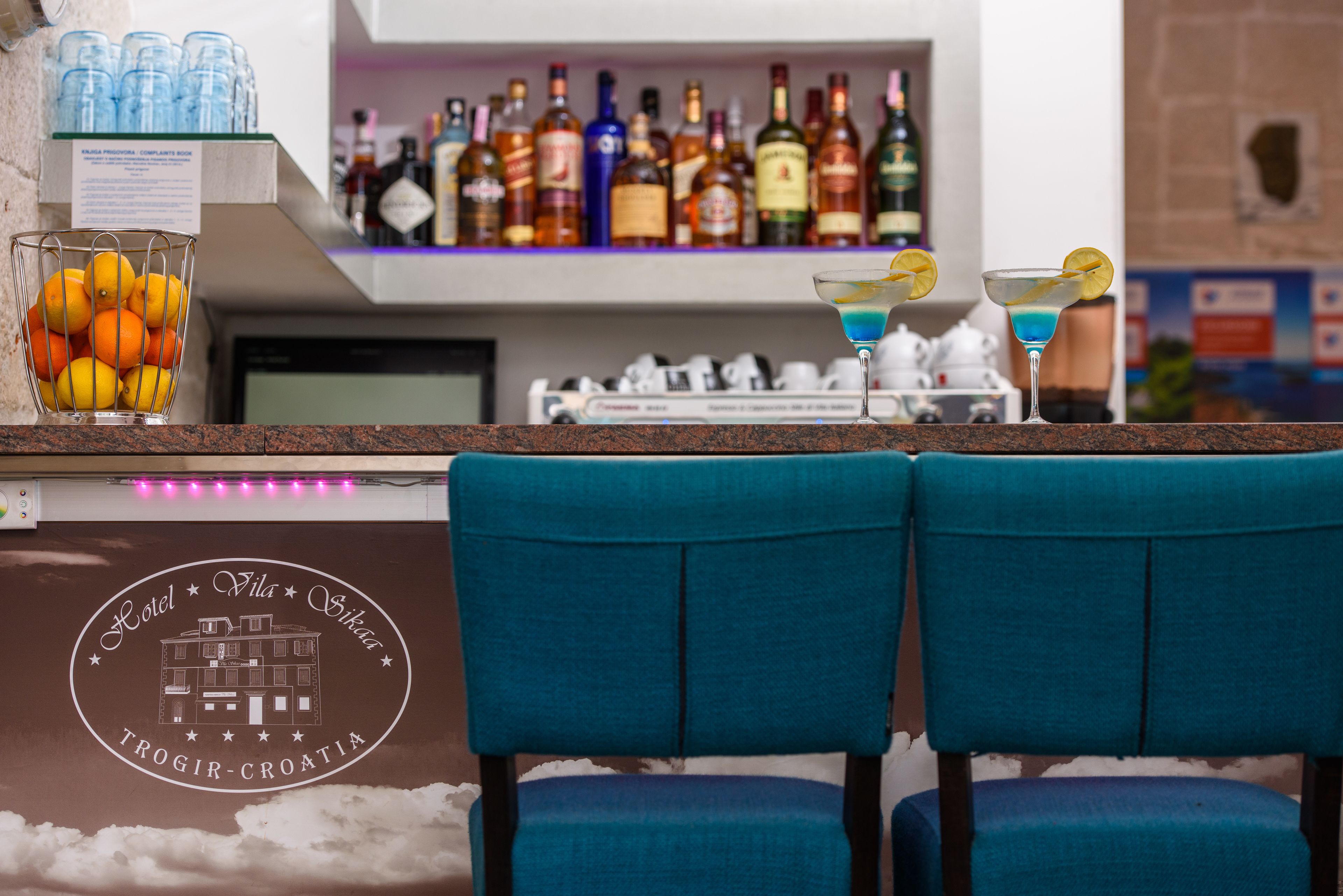 A bar area with a counter, a glass container of oranges, and two blue chairs.