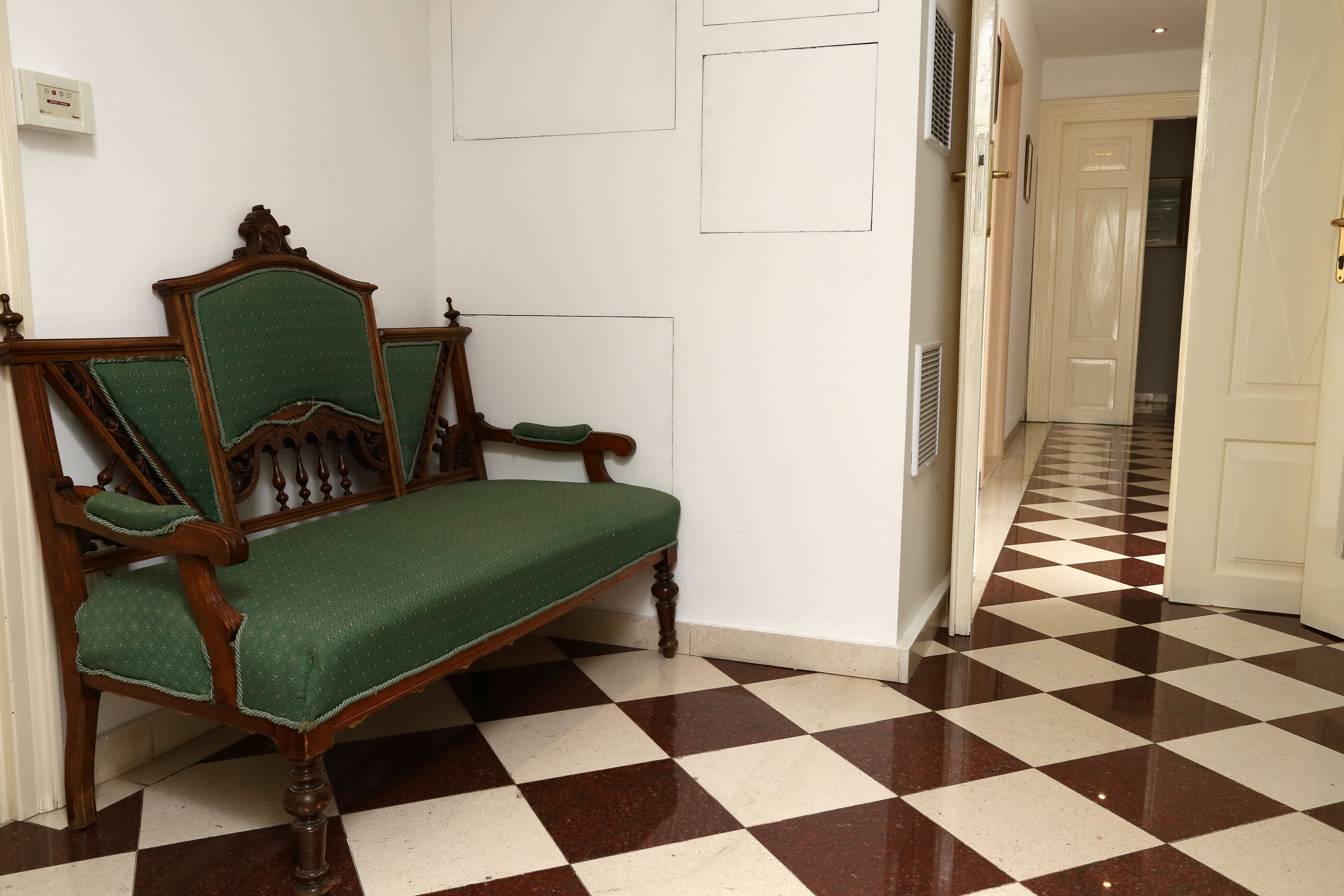 Una habitación con piso a cuadros, un banco de madera con cojín verde y una puerta blanca al fondo.