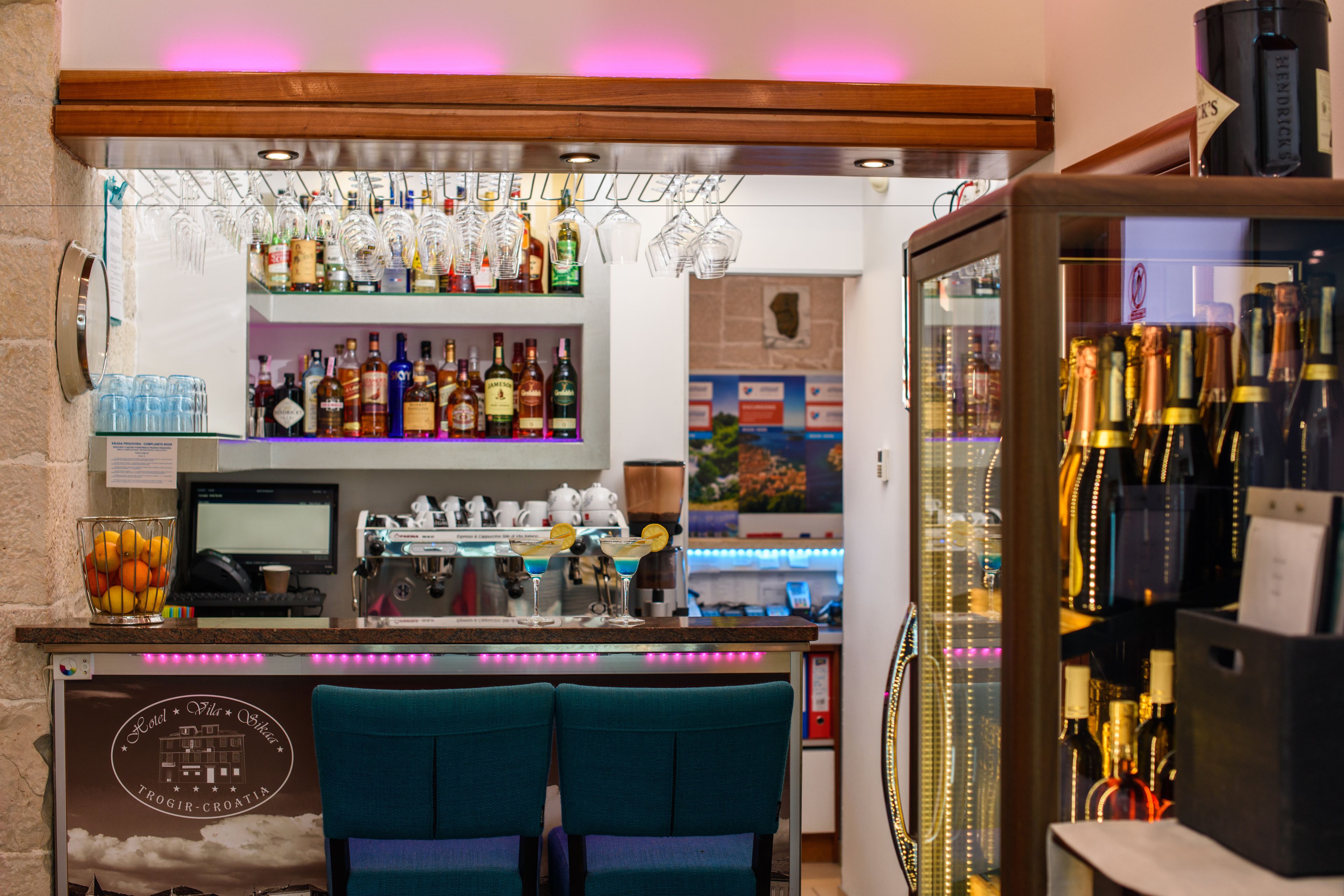 Una zona de bar con mostrador, taburetes y estantes llenos de botellas.