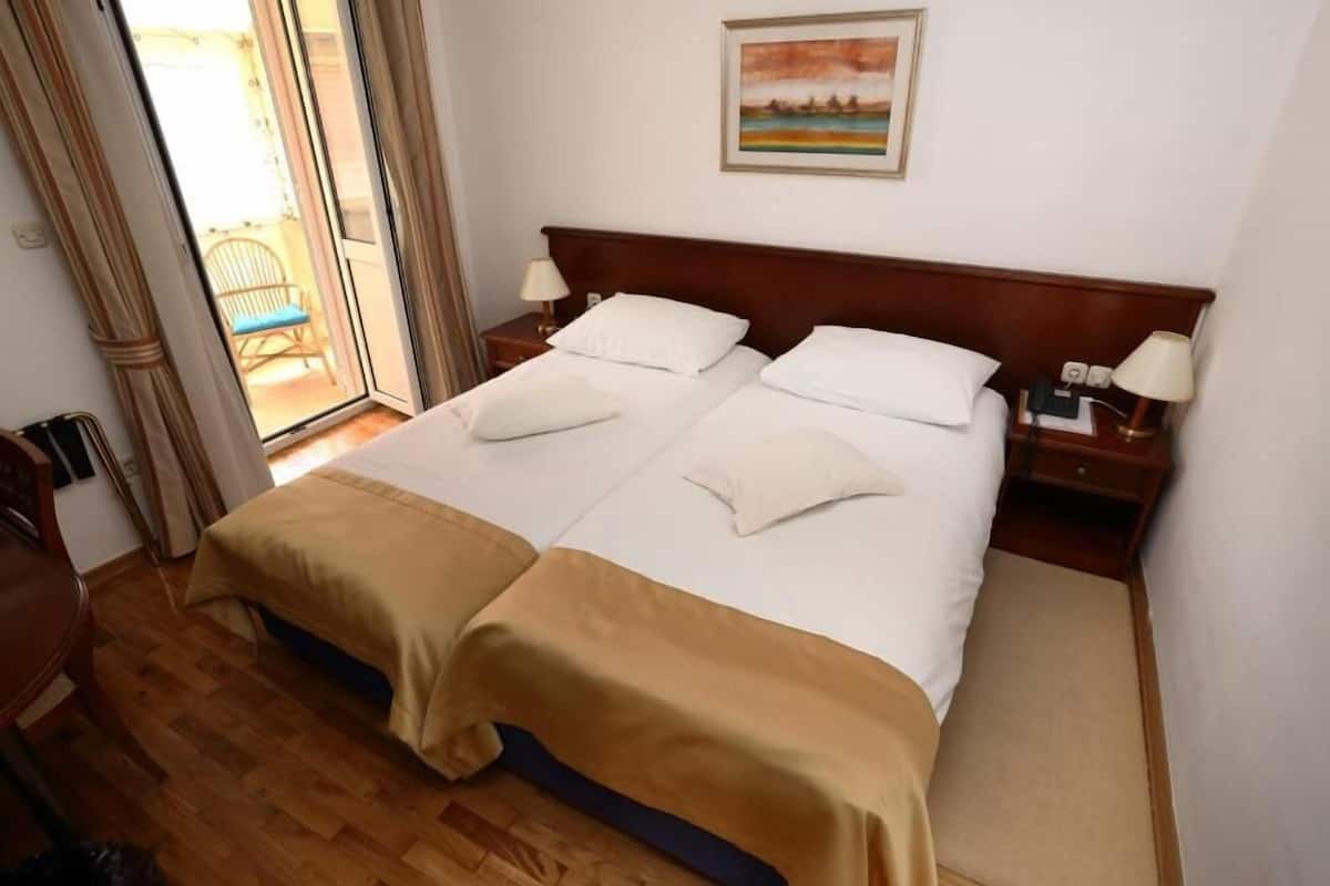 A hotel room with a double bed, wooden floor, two bedside tables with lamps, a painting on the wall, and a sliding glass door to the outside.