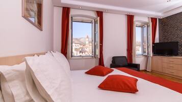 Habitación de hotel con una cama grande, dos almohadas rojas, un televisor, una silla y vistas de un paisaje urbano a través de las ventanas.