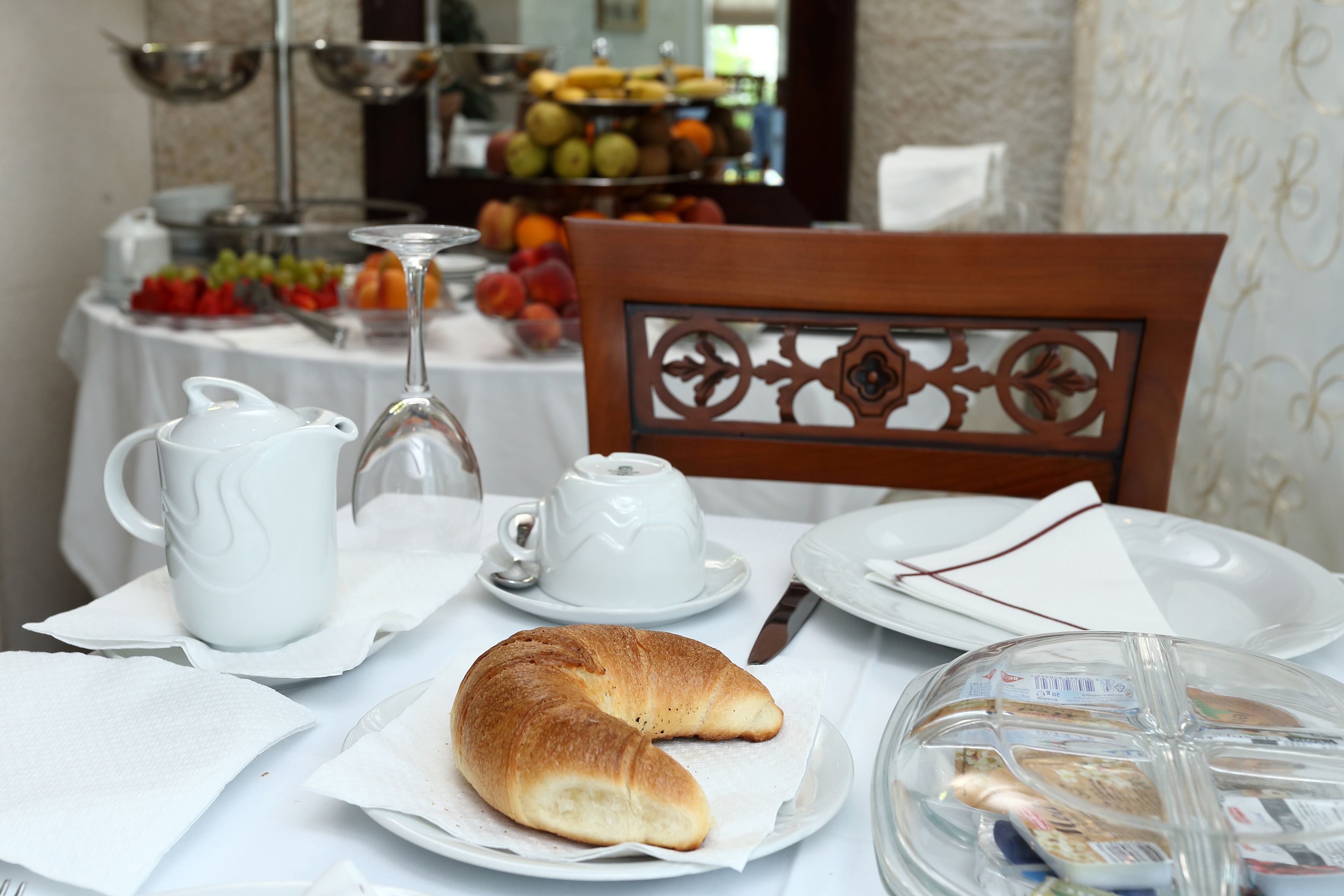 Un desayuno con croissants, tazas de café y una bandeja de frutas al fondo.