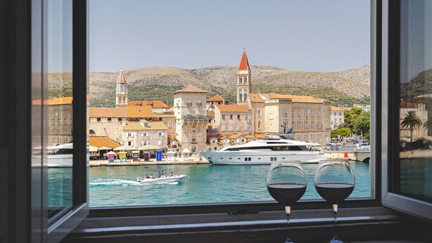 A view from a window overlooking a coastal town with historic buildings, a prominent tower, and a yacht docked in the water. - Hotel Sikaa (Trogir)