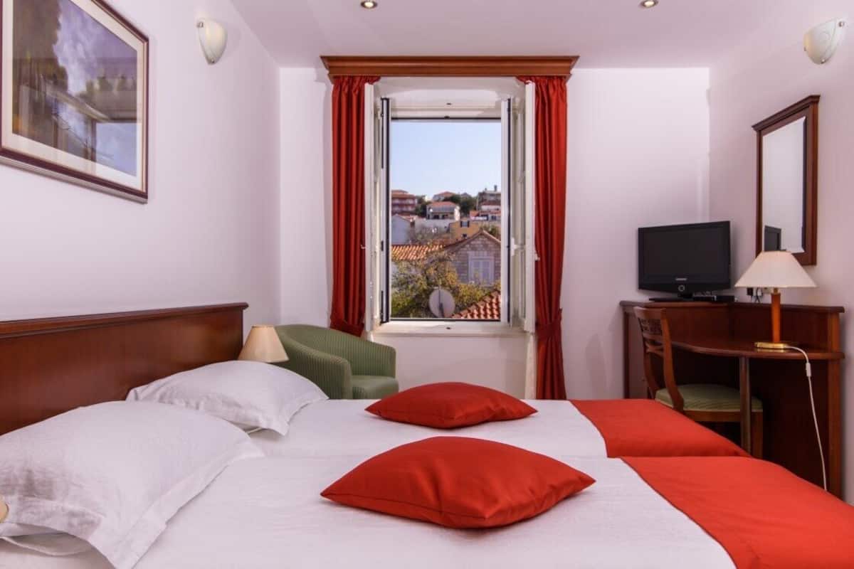 A hotel room with a large bed, a television, a desk with a lamp, and a window with red curtains.