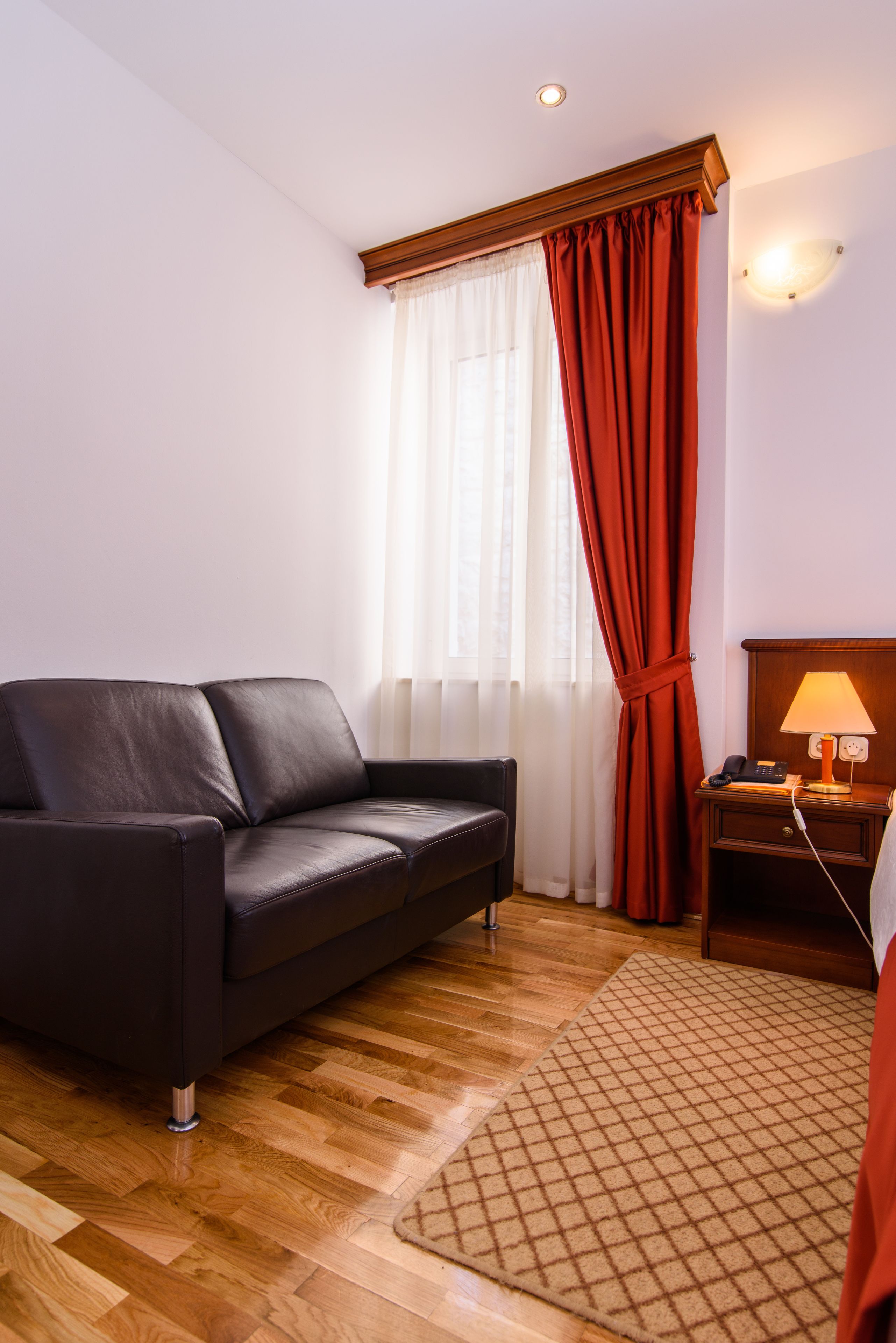 A modern hotel room with a leather sofa, wooden floor, a patterned rug, a nightstand with a lamp, and a window with red curtains.
