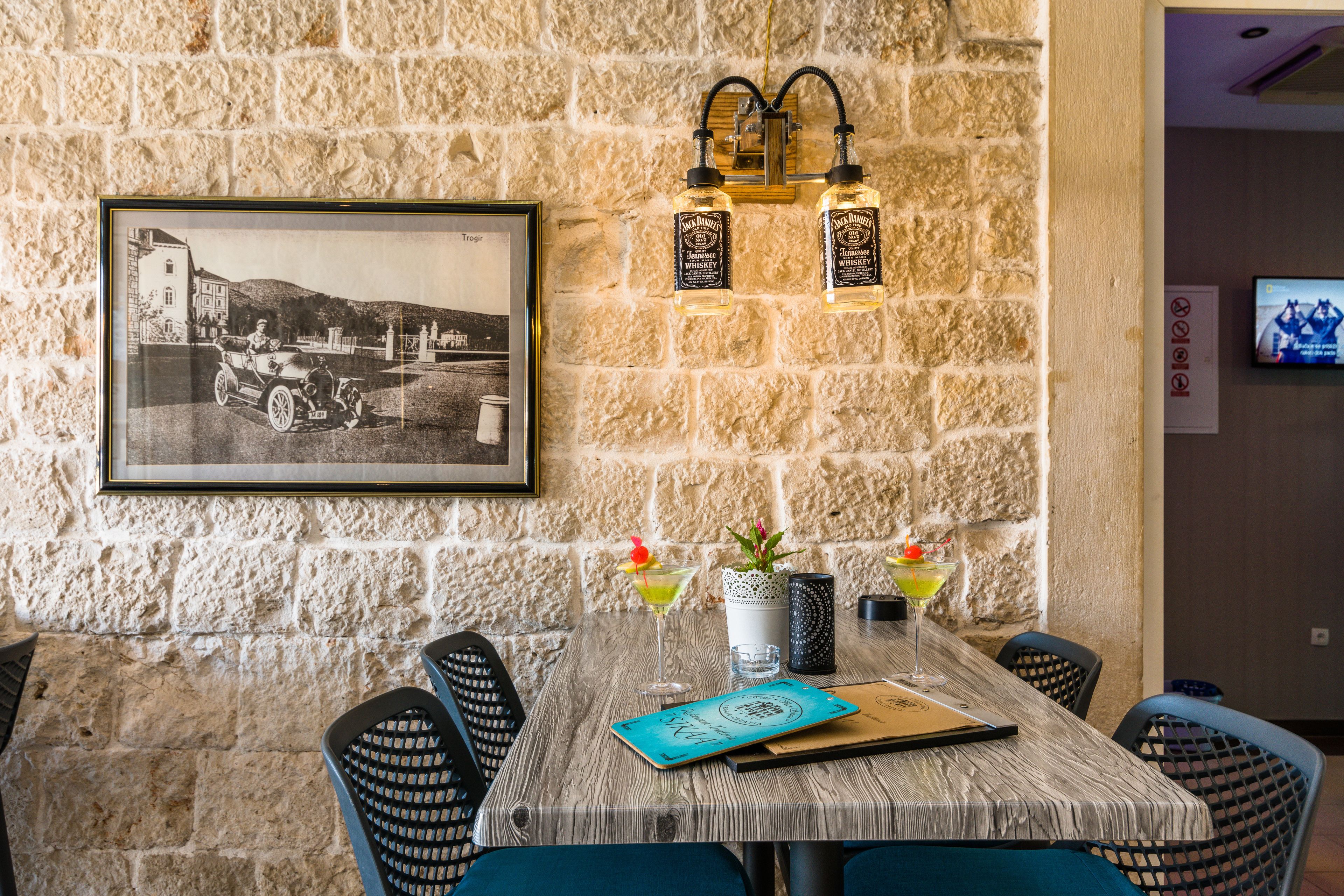 A rustic stone wall with a framed black and white photo, two whiskey bottles, a potted plant, and a menu on a wooden table with chairs.
