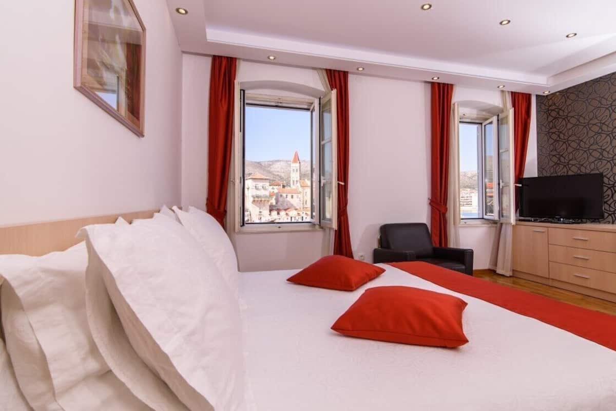 A hotel room with a bed, two red pillows, a TV, a chair, and a view of a cityscape through large windows.