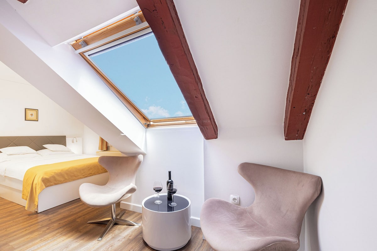 A modern bedroom with a skylight, a bed with a yellow blanket, a white chair, and a small table with glasses.