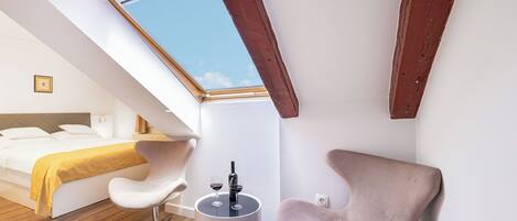 A modern bedroom with a skylight, a bed with a yellow blanket, a white chair, and a small table with glasses.