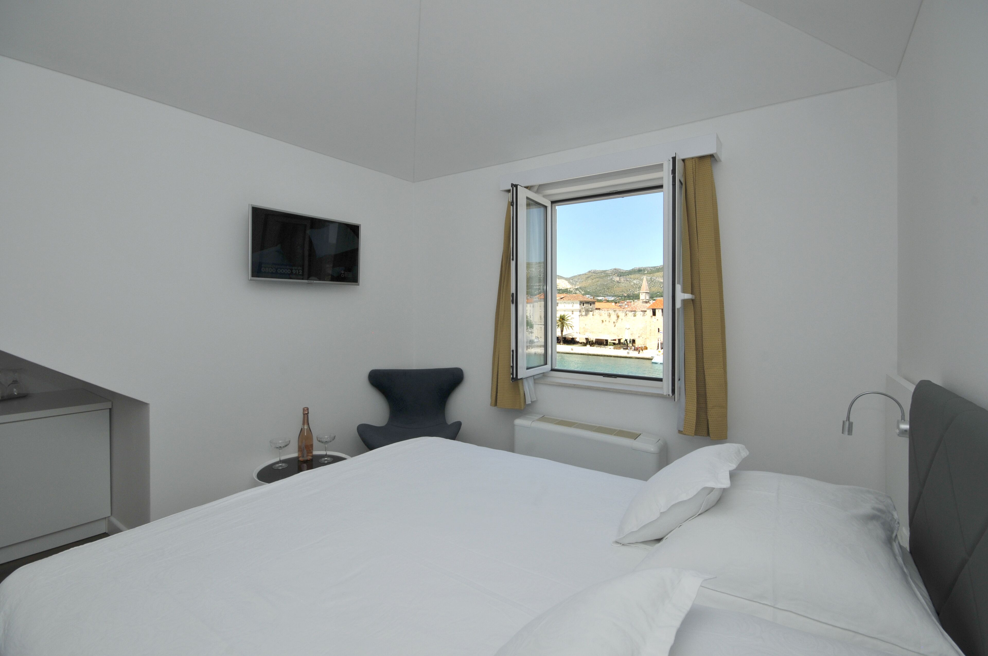A hotel room with a bed, a chair, a TV, and a window with a view of buildings and hills.