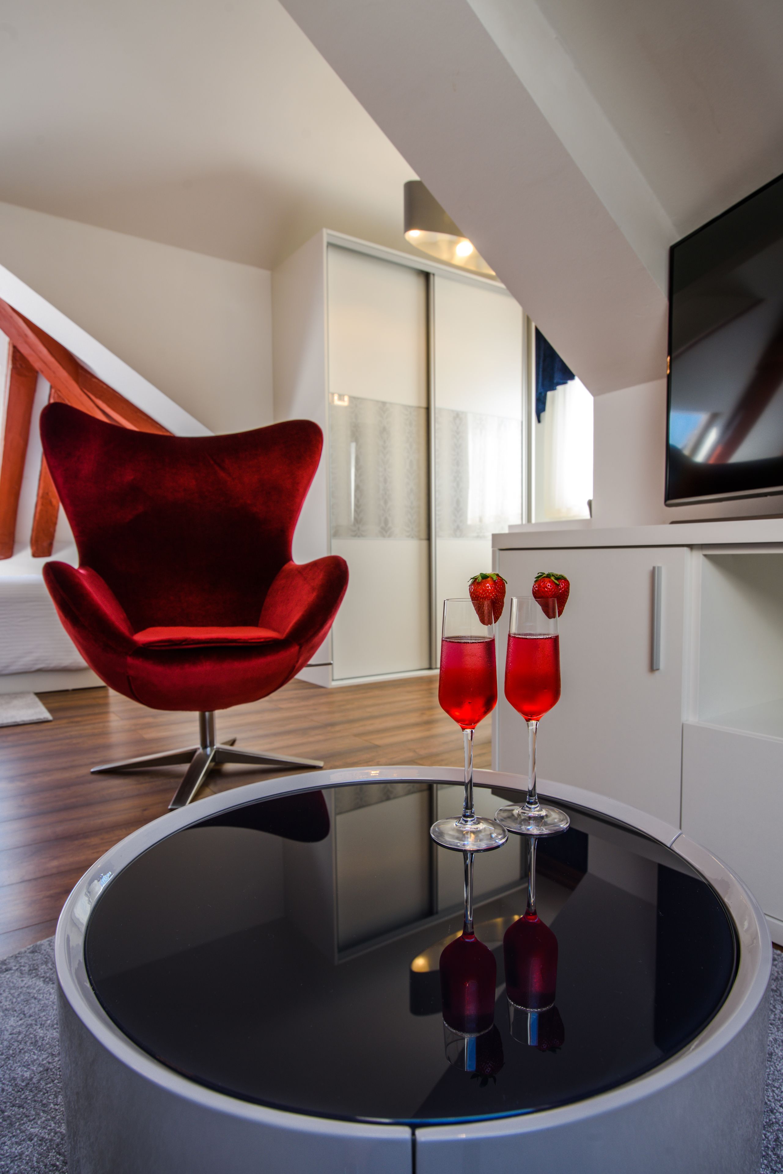 Ein modernes Wohnzimmer mit einem roten Samtstuhl, einem Glastisch mit zwei roten Getränken und Erdbeeren sowie einem Flachbildfernseher auf einem weißen Schrank.