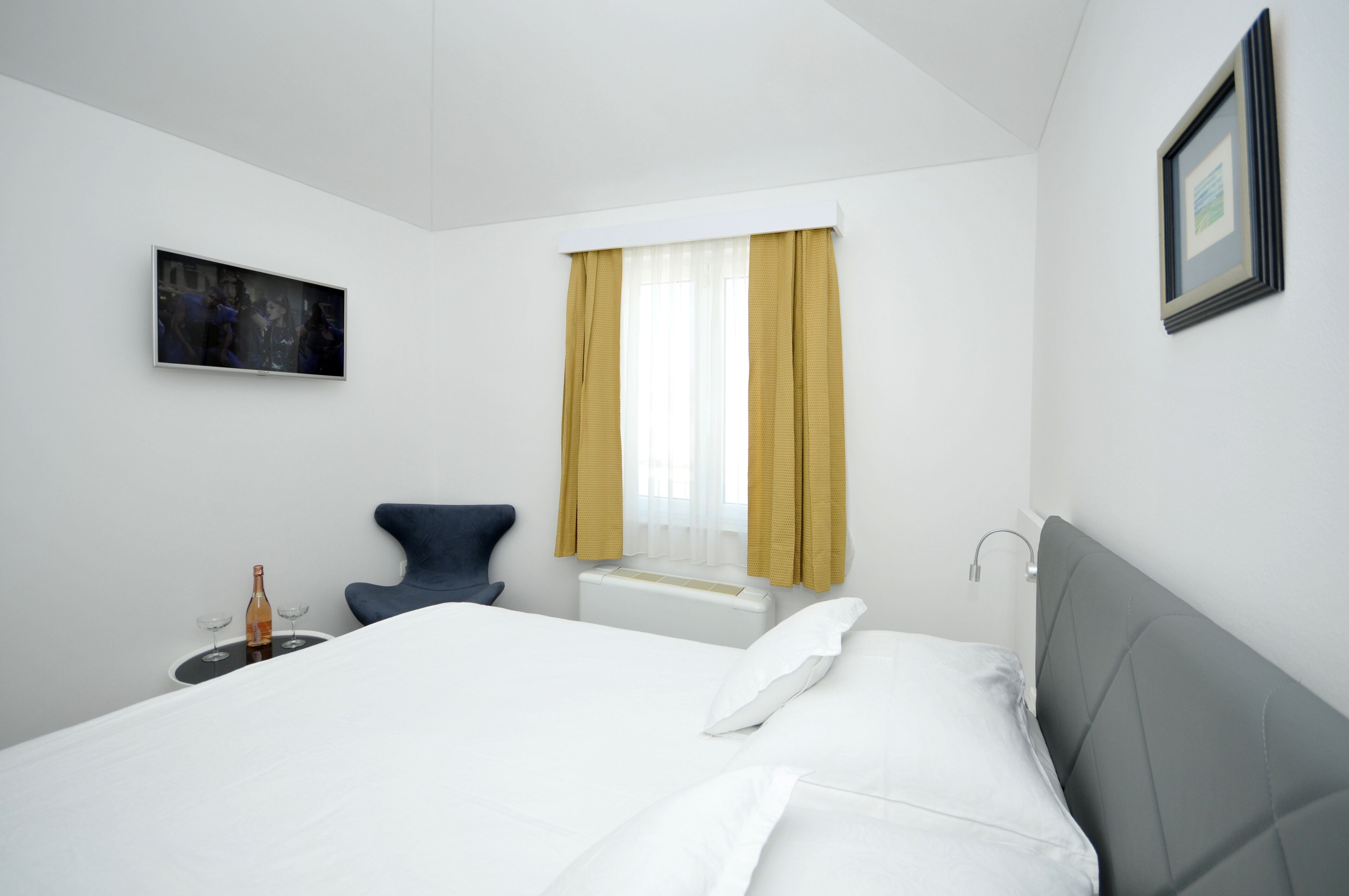 A hotel room with a bed, a chair, a small table with a bottle and glasses, a window with curtains, and a framed picture on the wall.