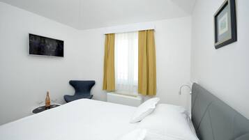 A hotel room with a bed, a chair, a small table with a bottle and glasses, a window with curtains, and a framed picture on the wall.